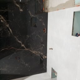 Usługi Ogólnobudowlane Marcin Mąka - Częściowo wykończona łazienka z czarnymi płytkami imitującymi marmur na jednej ścianie i białymi na drugiej, widoczne otwory na instalacje i brakujące fragmenty płytek przy podłodze.