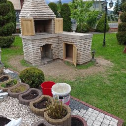 Usługi Ogólnobudowlane Marcin Mąka - Murowany grill ogrodowy z cegły szamotowej i zwykłej, z drewnianymi drzwiczkami, zlokalizowany na płycie fundamentowej, otoczony zielenią i elementami dekoracyjnymi ogrodu.