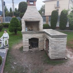 Usługi Ogólnobudowlane Marcin Mąka - Murowany grill z jasnej cegły z dodatkowym blatem roboczym, w Limanowej, obok rzeźby góralskiej z psem, na tle domu i zieleni.