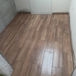 Usługi Ogólnobudowlane Marcin Mąka - Ułożone, brązowe płytki podłogowe imitujące drewno w małej pralni, widoczny fragment ściany z białą klapą rewizyjną oraz niedokończone obrzeże przy progu.