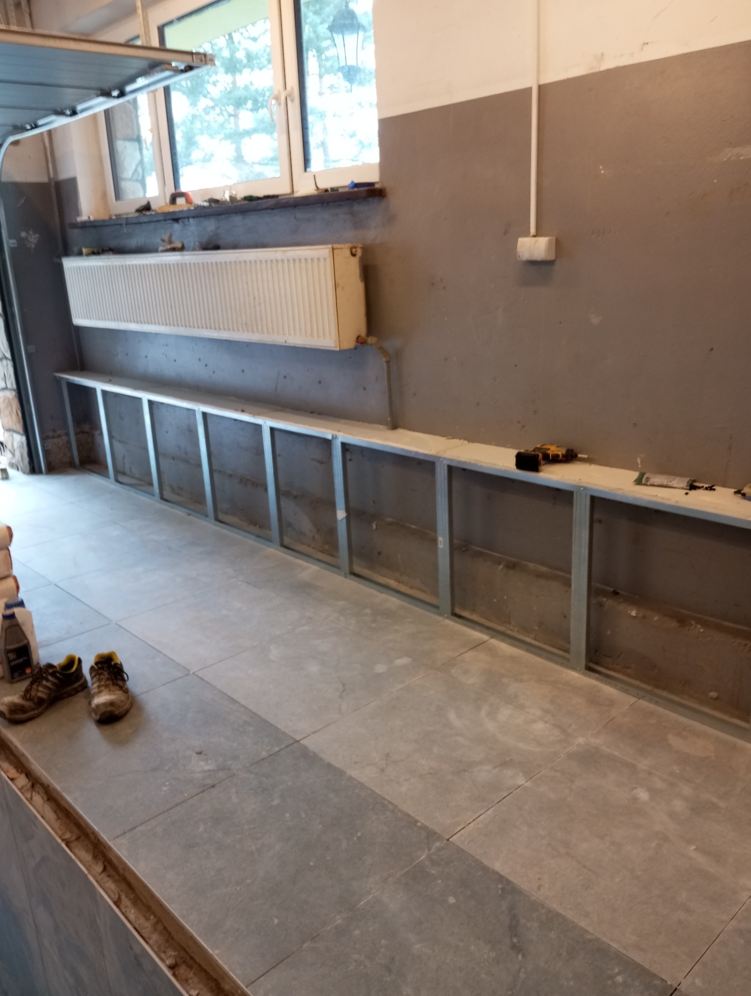Wnętrze w trakcie prac remontowych: ułożona szara podłoga z płytek, metalowa konstrukcja pod zabudowę ściany, grzejnik na ścianie, narzędzia pracy i buty robocze w rogu.