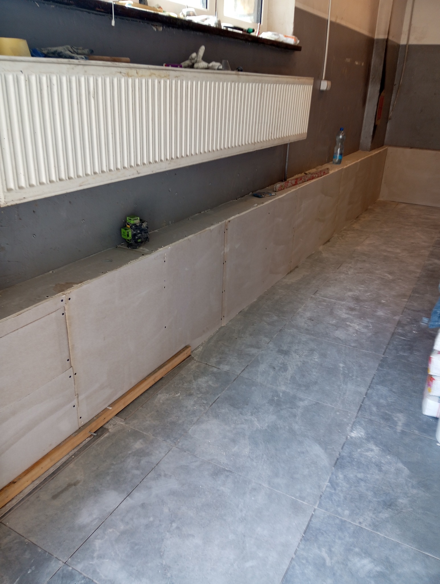 Wnętrze w trakcie prac: zabudowa z płyt karton-gips przy grzejniku, z widocznym laserowym niwelatorem na górnej krawędzi i fragmentem ułożonej podłogi z szarych płytek.