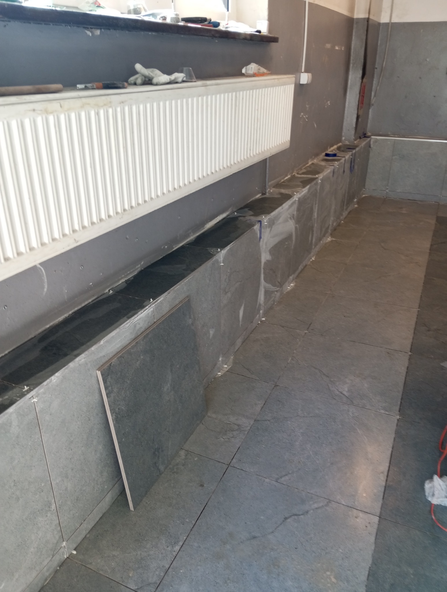 Szara płytka podłogowa i ścienna w trakcie układania, widoczne krzyżyki dystansowe, fragment grzejnika i narzędzia na półce powyżej.