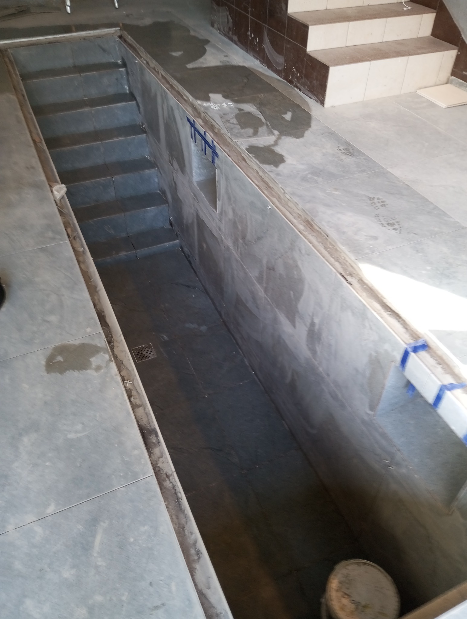 Wykładanie schodów i podłogi płytkami ceramicznymi w nowo wykończonym pomieszczeniu, widoczny odpływ liniowy w podłodze, zabezpieczenia na krawędziach płytek.