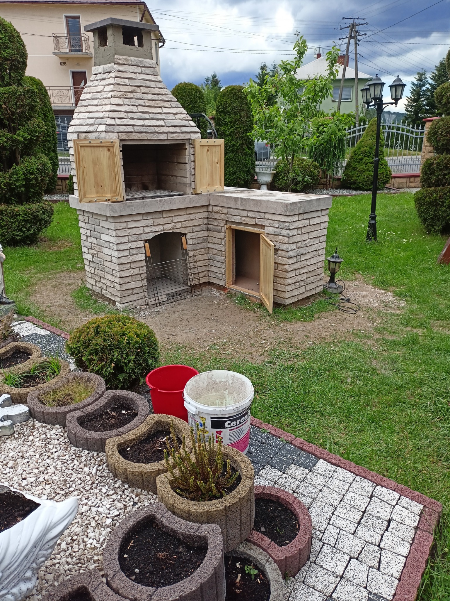 Murowany grill ogrodowy z cegły szamotowej i zwykłej, z drewnianymi drzwiczkami, zlokalizowany na płycie fundamentowej, otoczony zielenią i elementami dekoracyjnymi ogrodu.