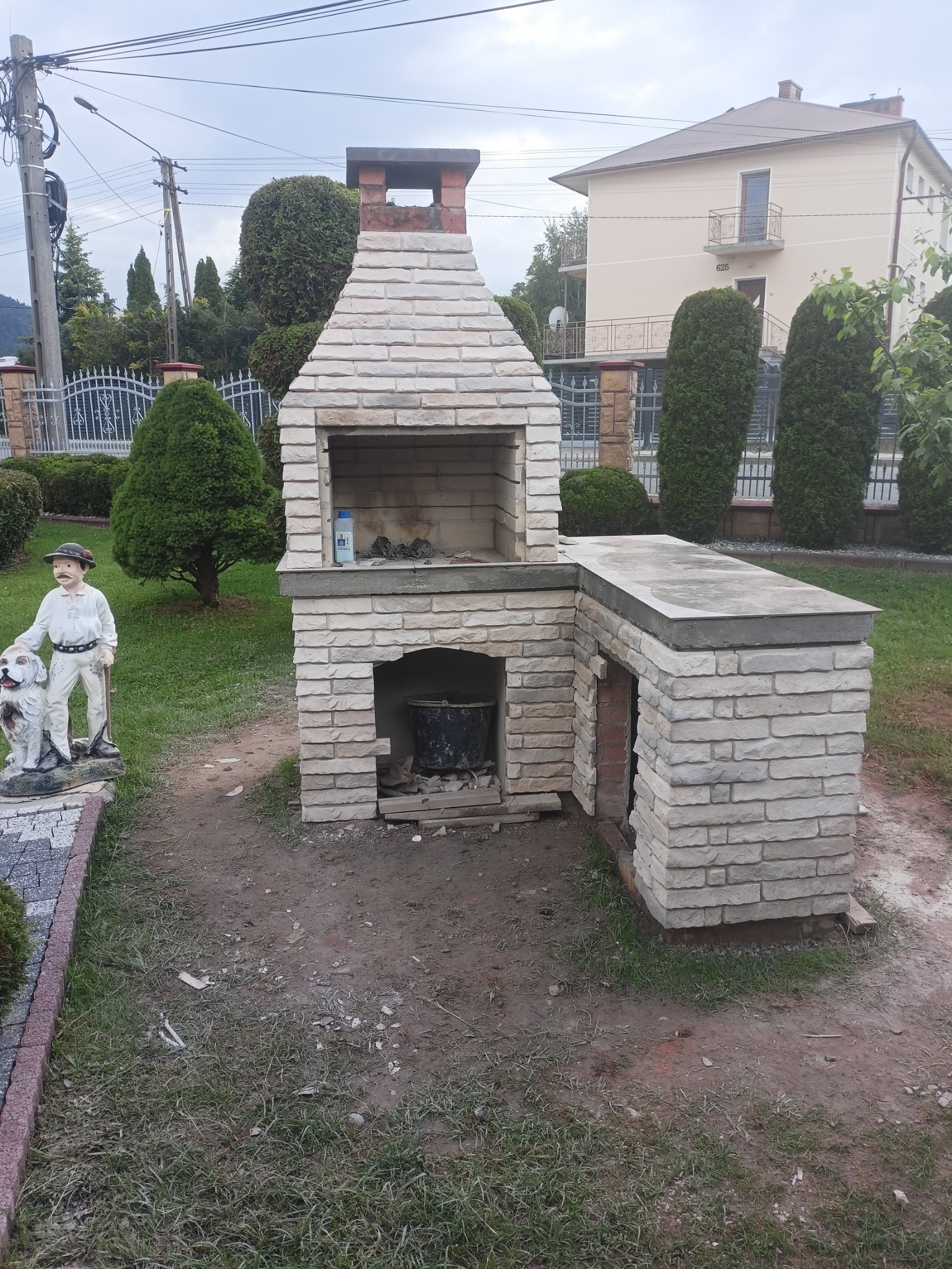 Murowany grill z jasnej cegły z dodatkowym blatem roboczym, w Limanowej, obok rzeźby góralskiej z psem, na tle domu i zieleni.