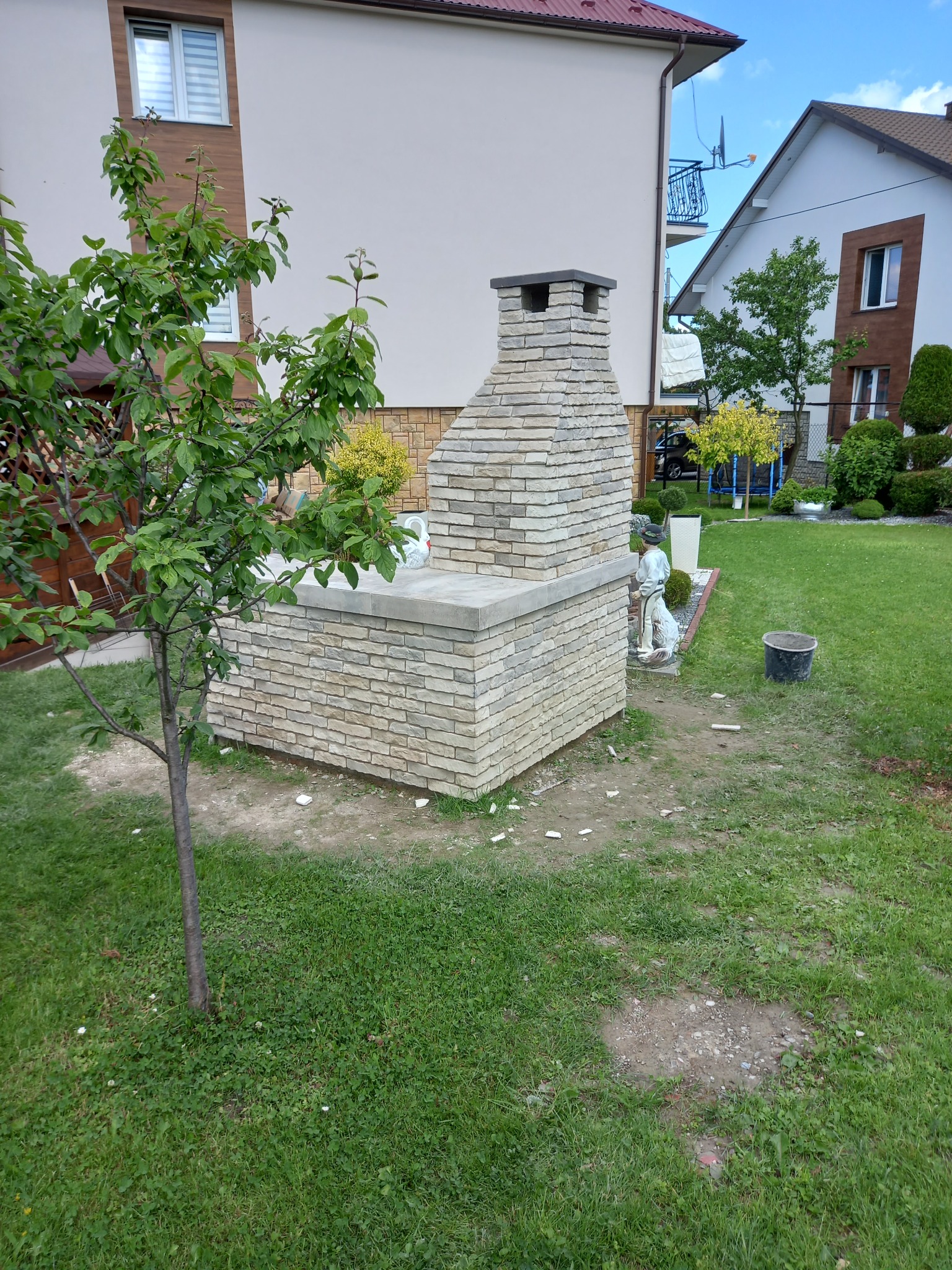 Murowany grill ogrodowy z jasnej cegły, z kominem, na trawniku przed domem, obok stoi ozdobna figurka chłopca.