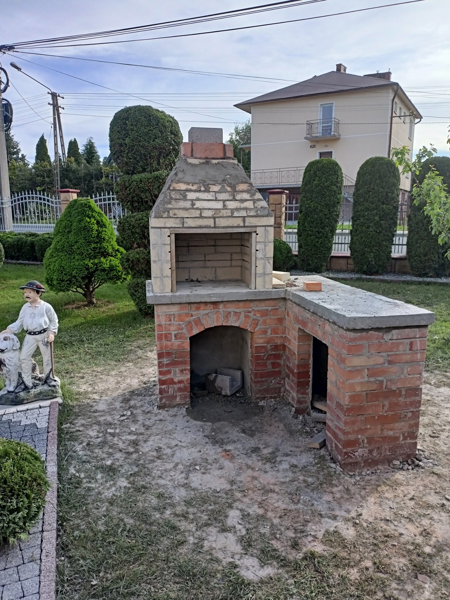 Murowany grill ogrodowy z cegły, w trakcie budowy, widoczny na tle zadbanego ogrodu i domu, obok dekoracyjna figurka górala z psem.