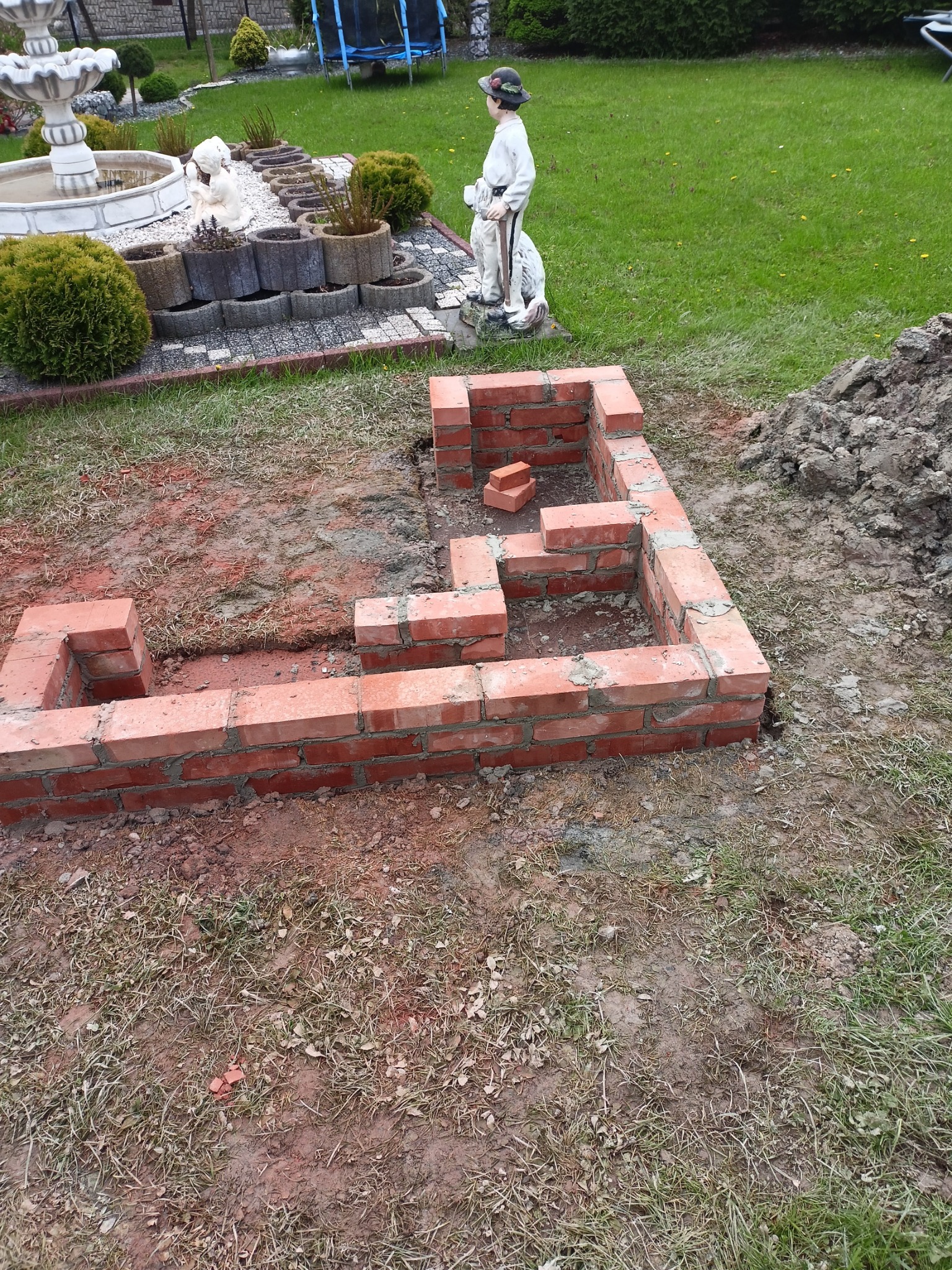 Fundament z czerwonej cegły, w trakcie budowy, widoczny na trawniku w ogrodzie, z fontanną i figurką w tle.