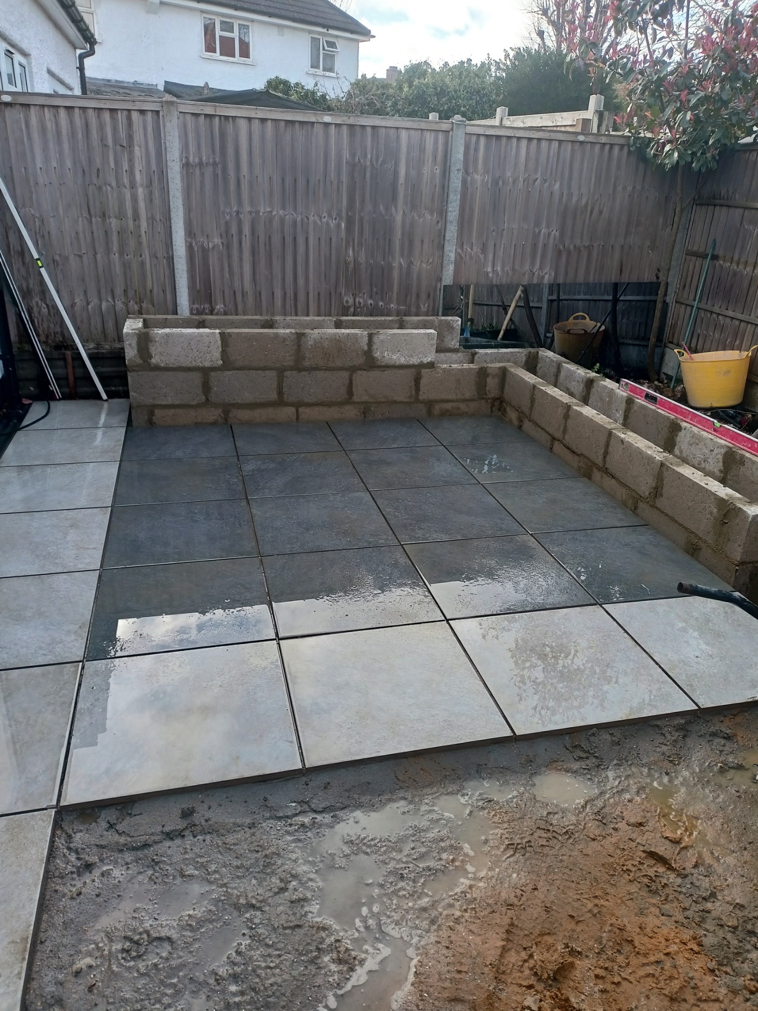 Wykładanie tarasu płytkami gresowymi w ogrodzie, widoczne ułożone płytki, przygotowane podłoże, murki z bloczków betonowych i drewniane ogrodzenie.