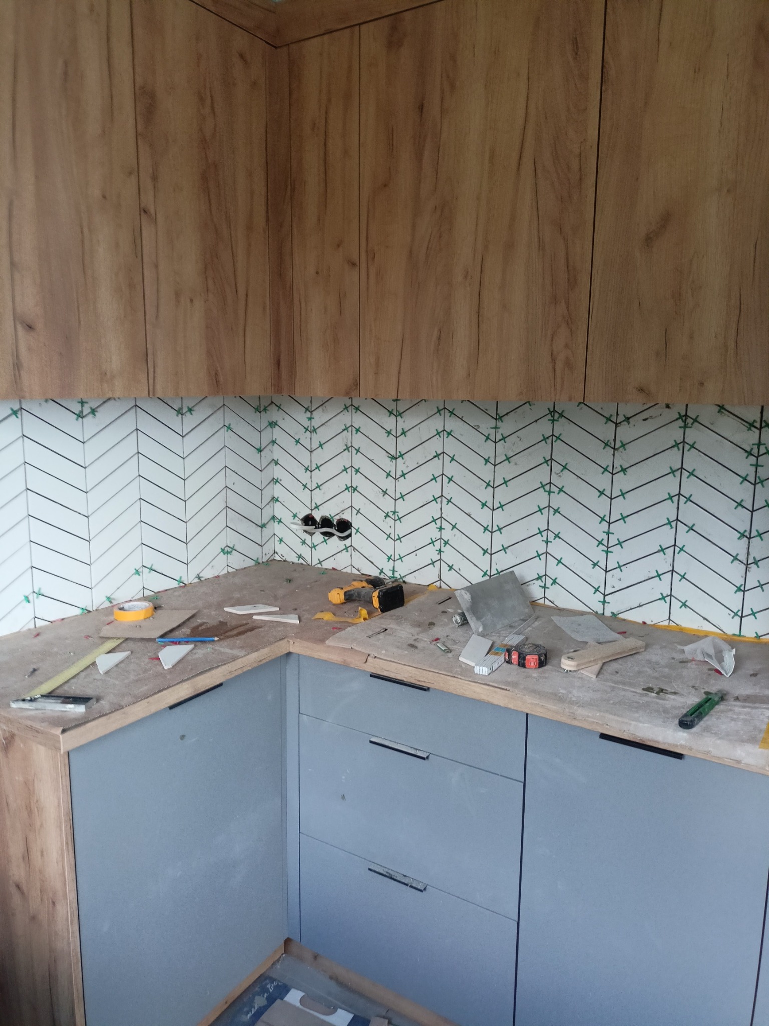 Montaż białych płytek ułożonych w jodełkę nad szarym blatem kuchennym, widoczne narzędzia i krzyżyki dystansowe.