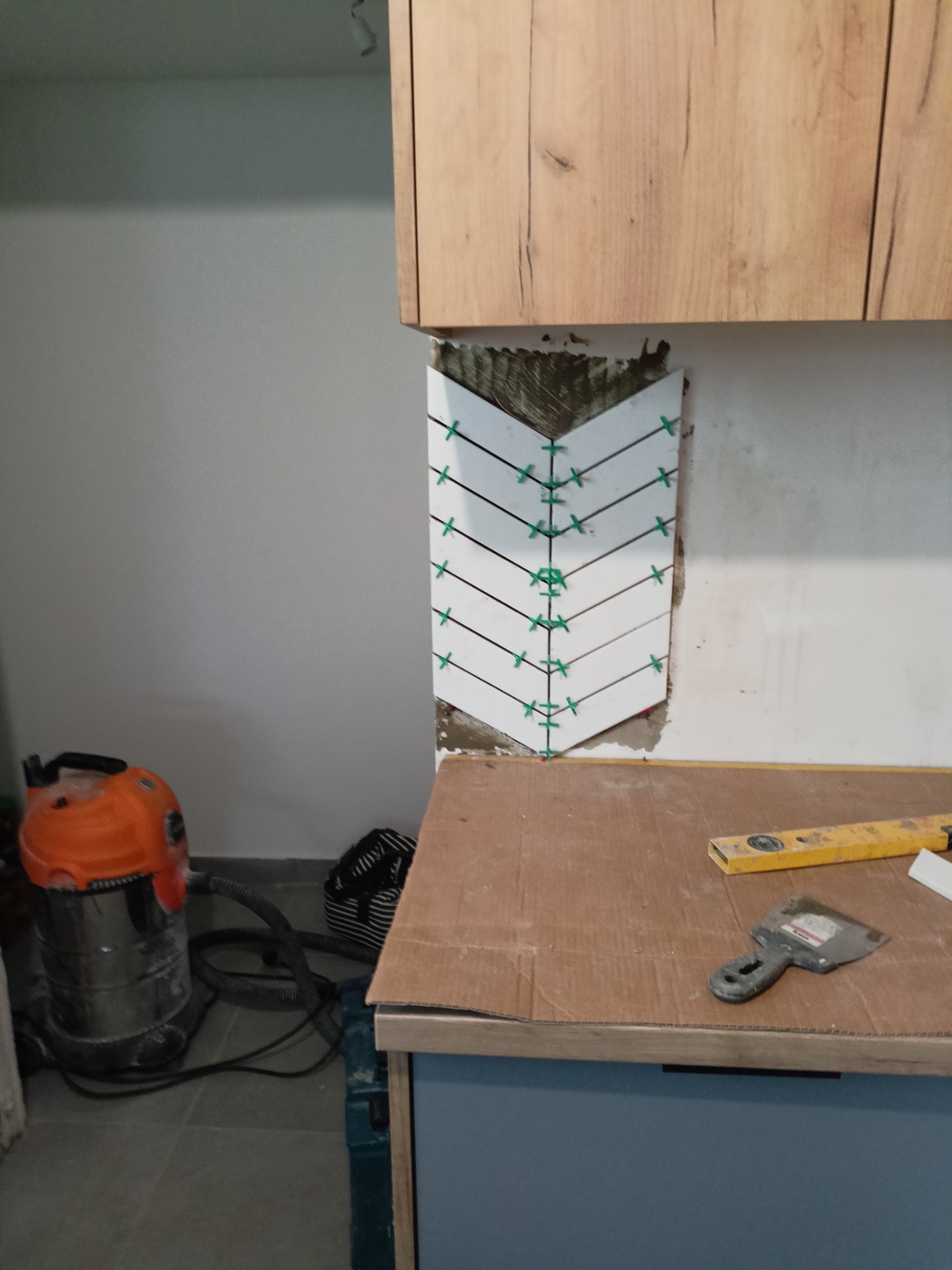 Białe płytki 3D ułożone w jodełkę na ścianie nad blatem kuchennym, z użyciem krzyżyków dystansowych, widoczny poziomica i kielnia na blacie oraz odkurzacz przemysłowy.