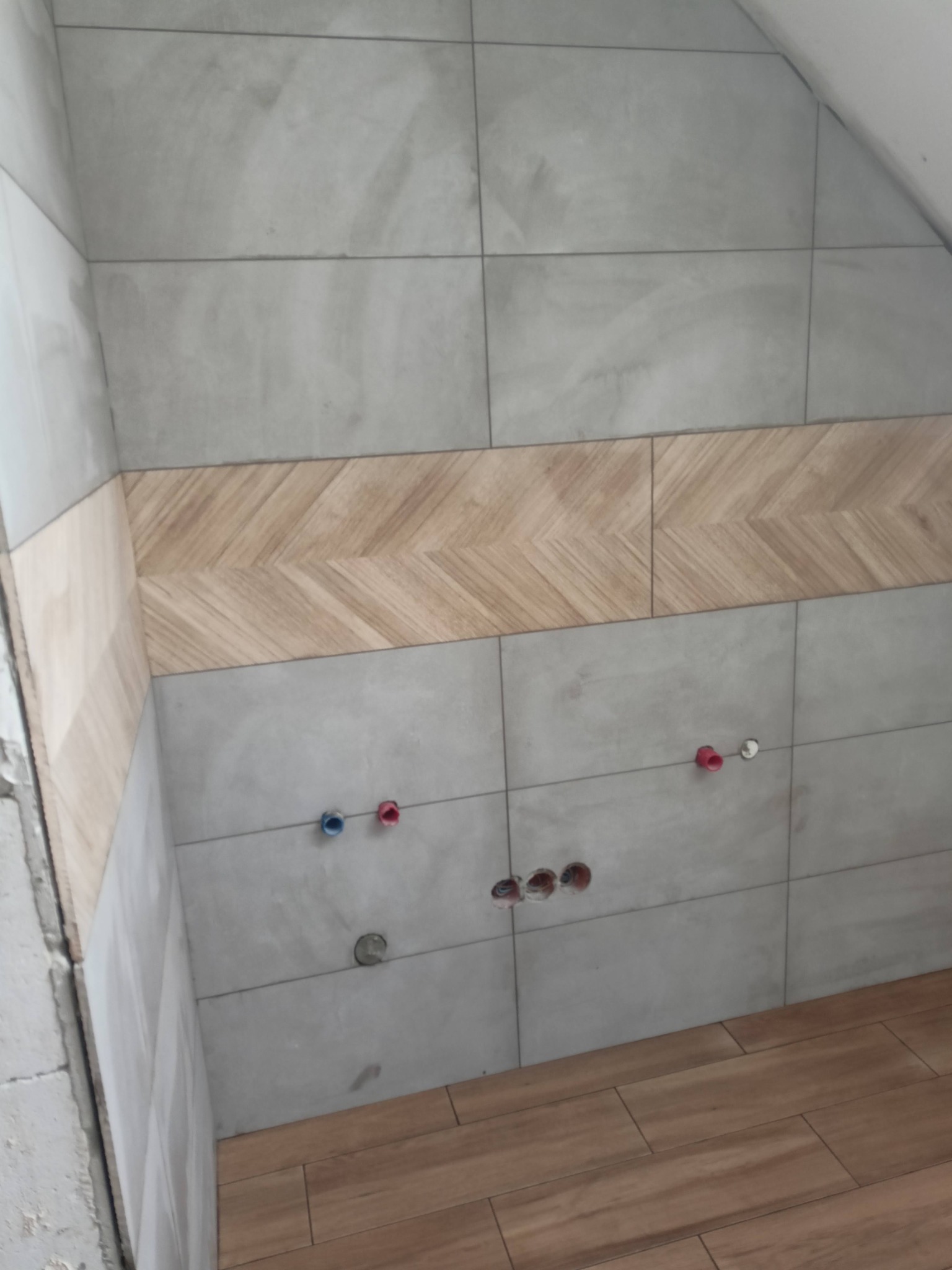 Fragment ściany w trakcie wykańczania, z szarymi płytkami i dekoracyjnym pasem z płytek imitujących drewno ułożonych w jodełkę, widoczne otwory na instalacje wodne i częściowo ułożona podłoga...