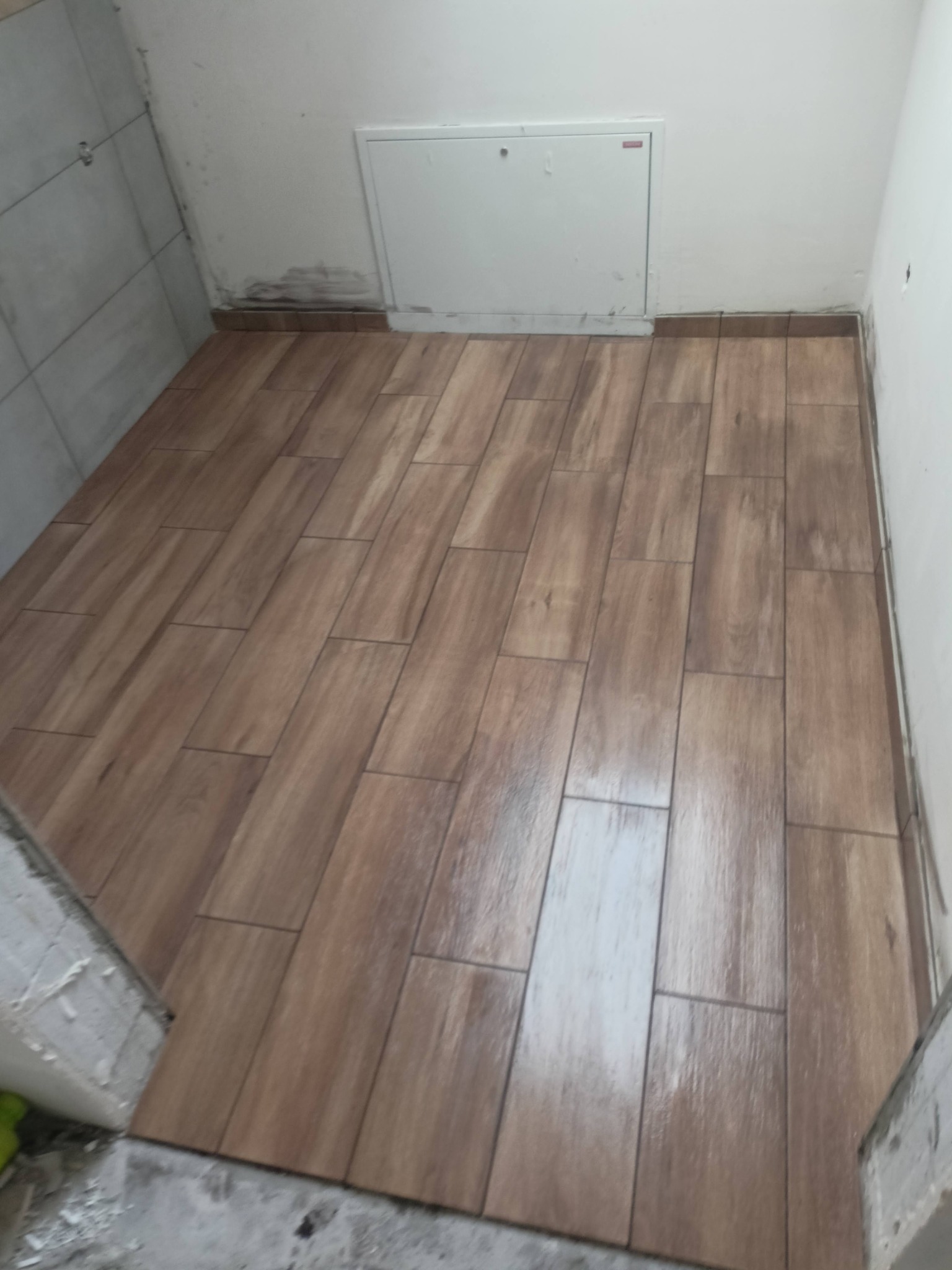 Ułożone, brązowe płytki podłogowe imitujące drewno w małej pralni, widoczny fragment ściany z białą klapą rewizyjną oraz niedokończone obrzeże przy progu.