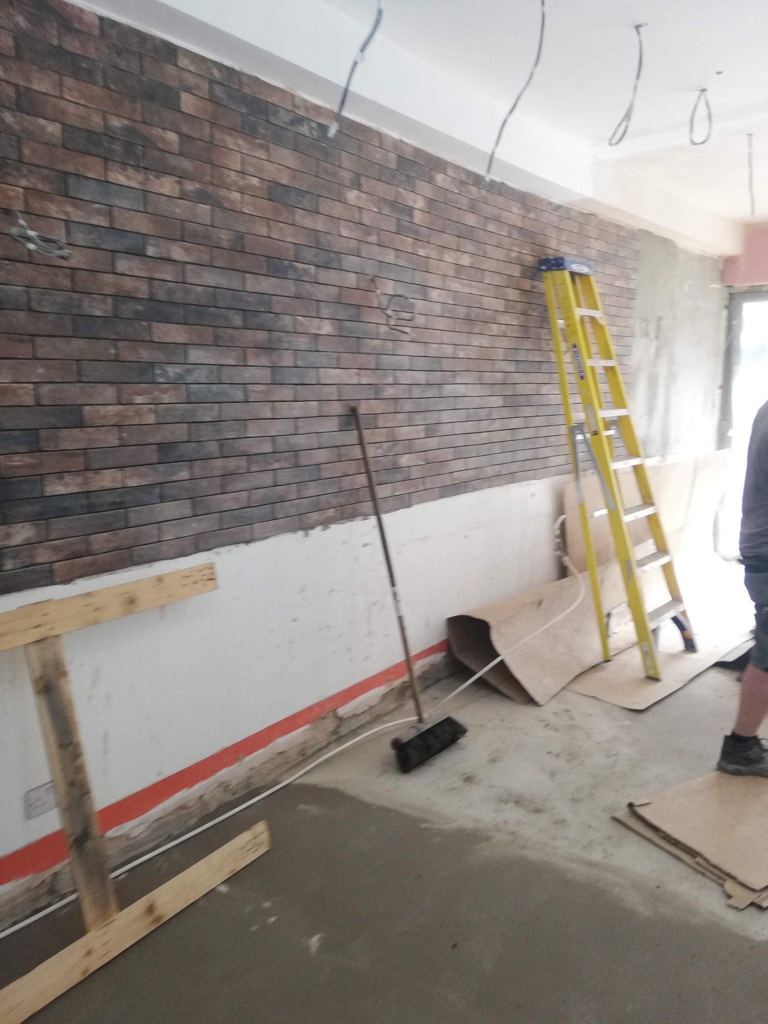 Ściana wykończona panelami imitującymi cegłę w trakcie montażu, widoczna drabina, narzędzia i surowe wykończenie podłogi, przewody elektryczne wystające z sufitu.