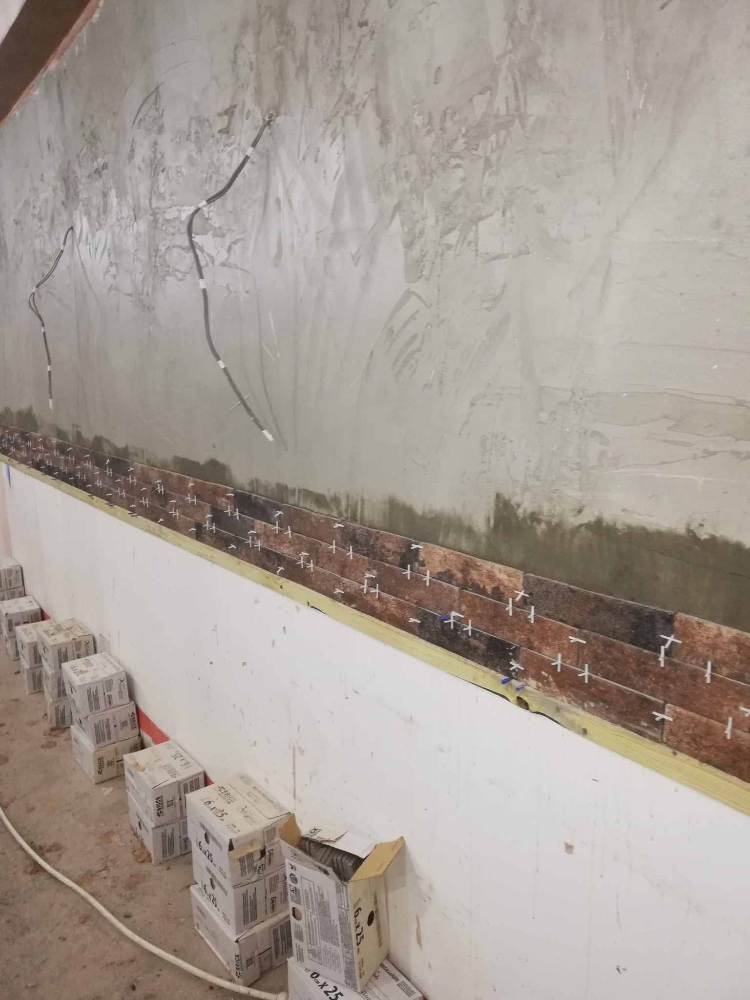 Ściana w trakcie wykańczania imitacją kamienia: widoczny fragment surowej ściany, rząd ułożonych płytek z krzyżykami dystansowymi i kartony płytek na podłodze.