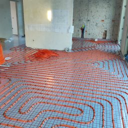 Hydra Justyna Radtke - Pomarańczowe rury ogrzewania podłogowego ułożone na srebrnej folii z siatką, widoczne fragmenty ścian w surowym stanie i instalacje elektryczne.