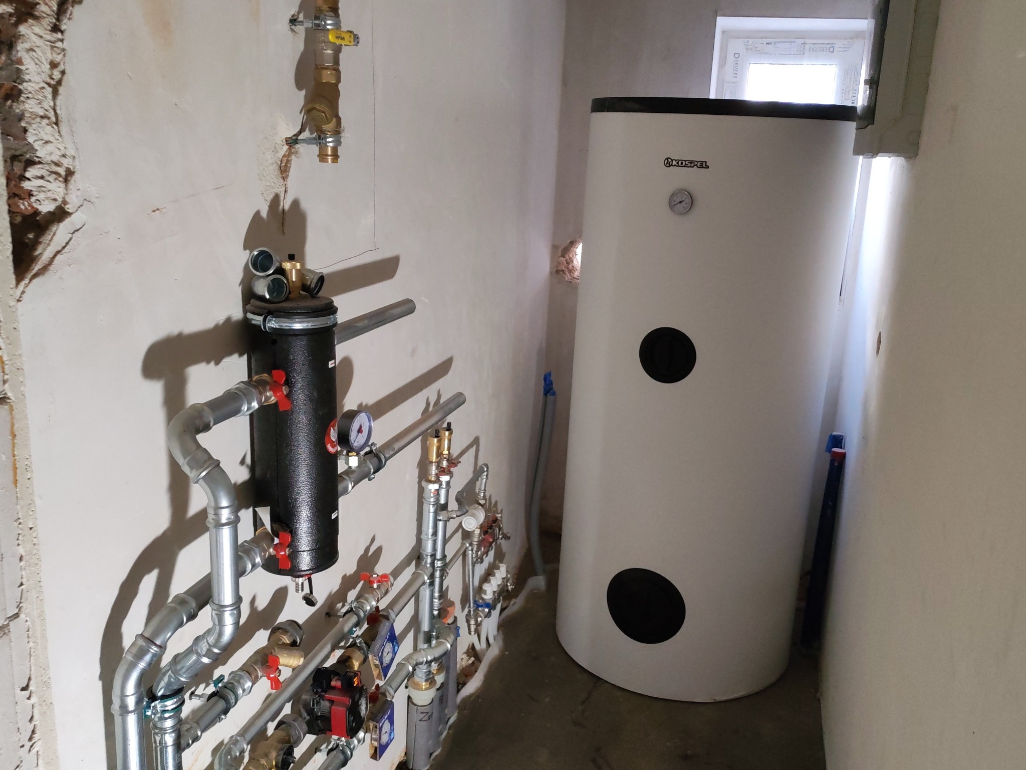 Biały bojler Kospel z czarnymi okręgami i instalacja hydrauliczną z zaworami w surowym pomieszczeniu z oknem. Widoczne rury i elementy montażowe.