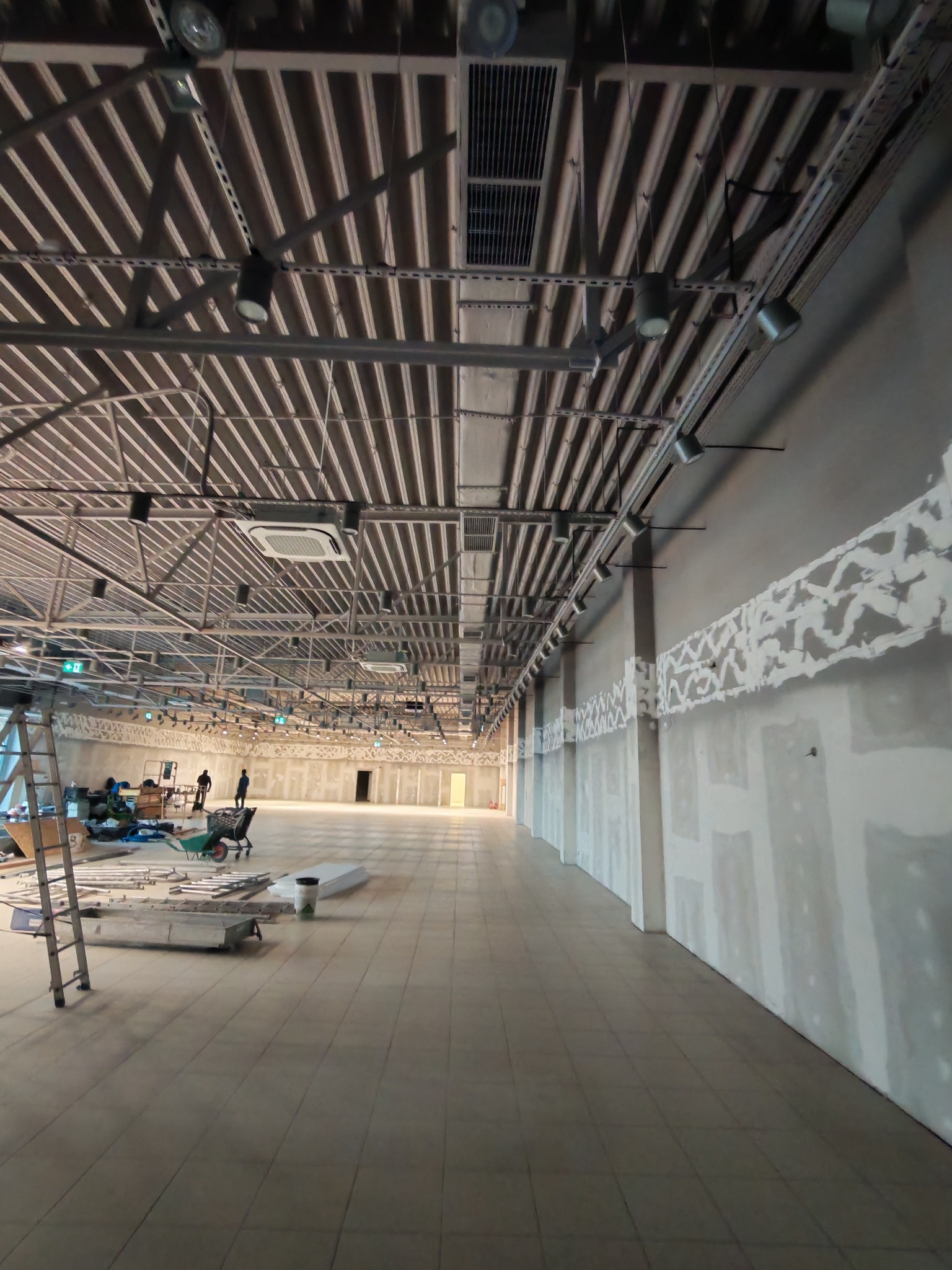 Wnętrze hali w trakcie prac wykończeniowych: widoczne instalacje pod sufitem, ściany z fragmentami gładzi, drabina i narzędzia na podłodze. Perspektywa z dołu.