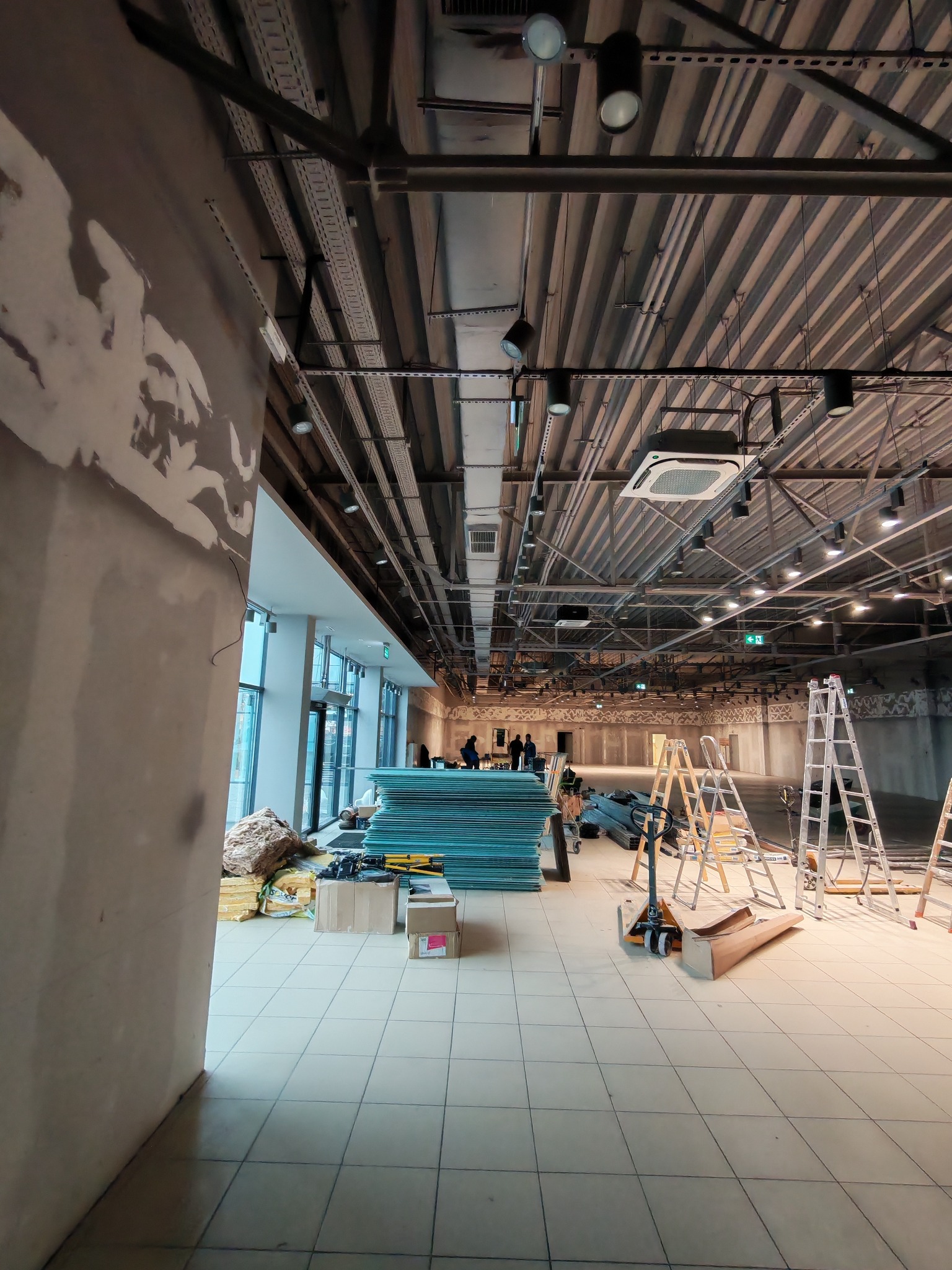 Wnętrze komercyjne w trakcie prac instalacyjnych: widoczne instalacje pod sufitem, materiały budowlane, drabiny i paleciak. Ściany w stanie surowym, przygotowane do wykończenia.