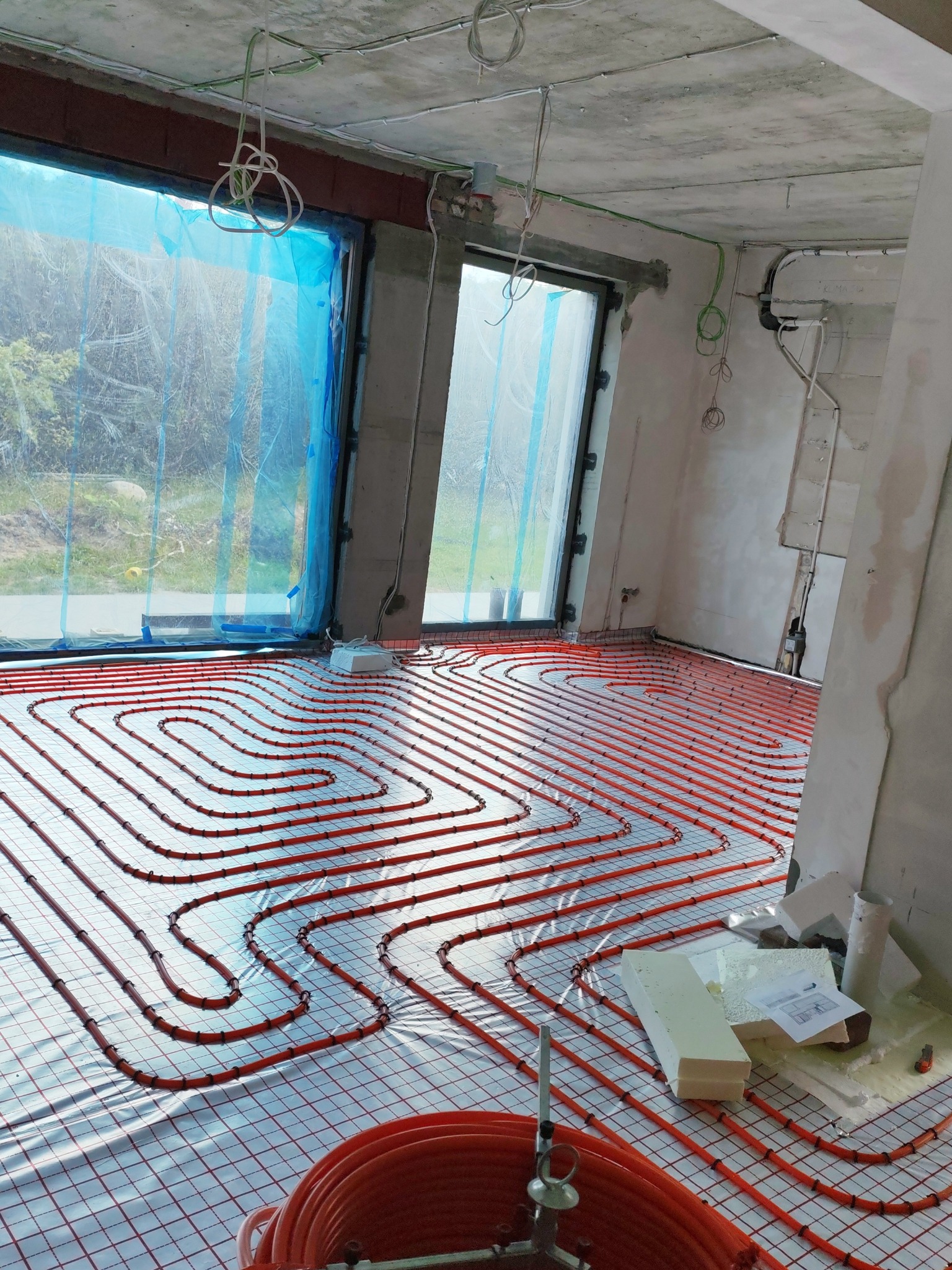 Pomarańczowe rury ogrzewania podłogowego ułożone na siatce w surowym wnętrzu. Okna zabezpieczone folią, widoczne przewody elektryczne na suficie.