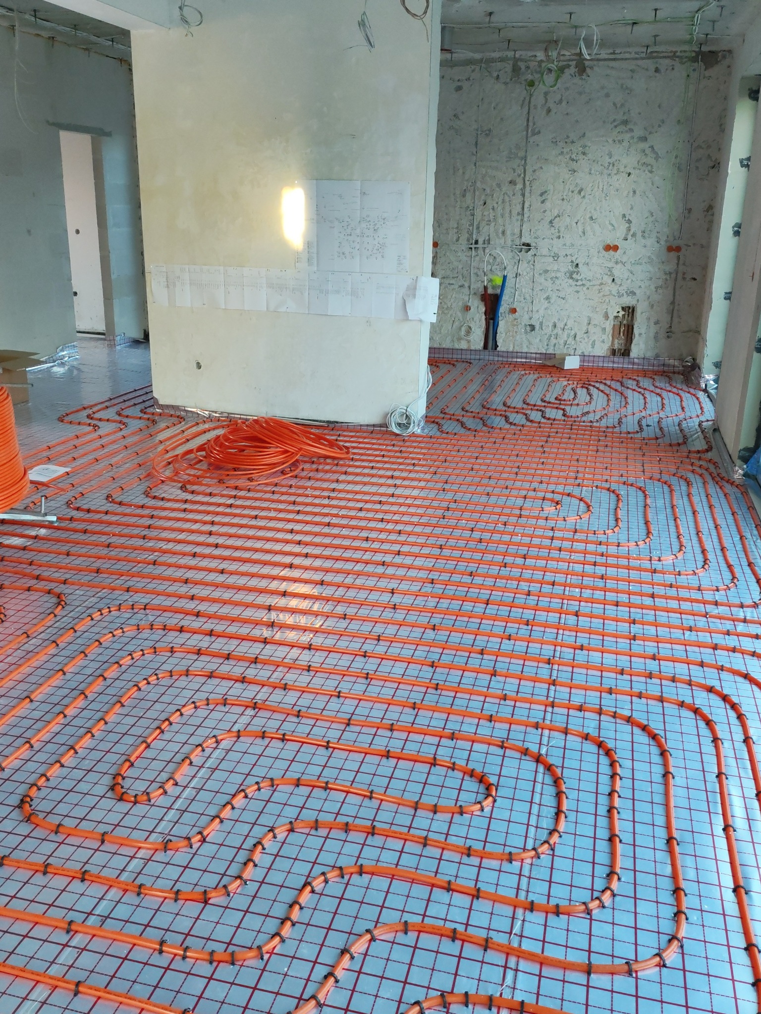 Pomarańczowe rury ogrzewania podłogowego ułożone na srebrnej folii z siatką, widoczne fragmenty ścian w surowym stanie i instalacje elektryczne.