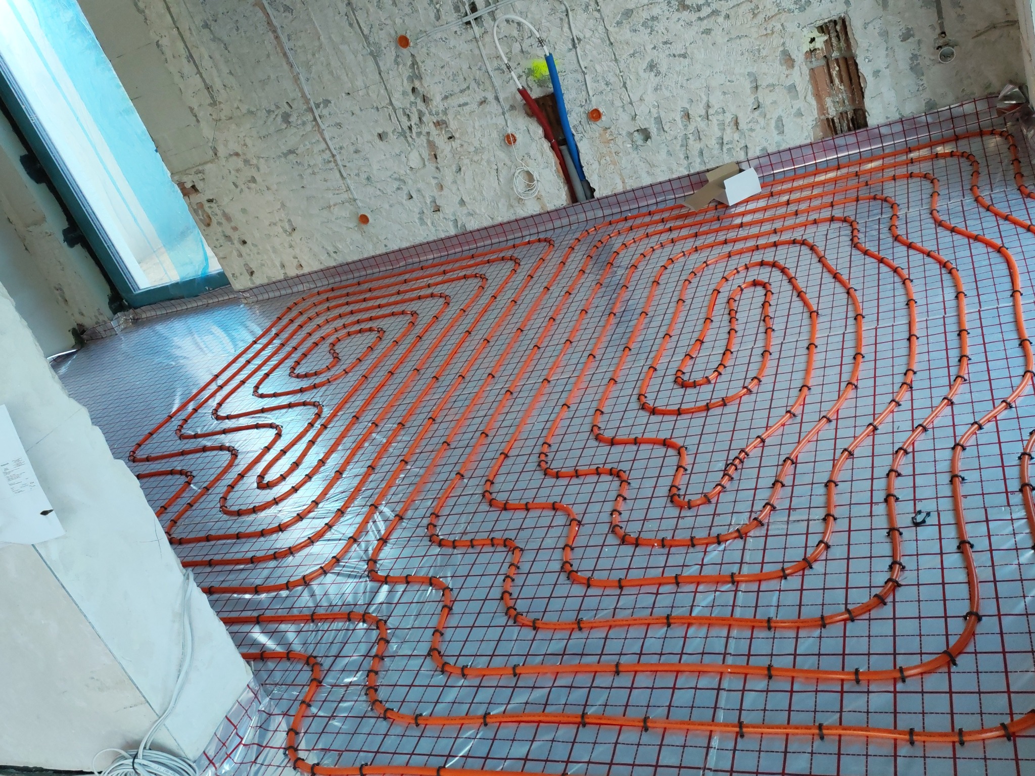 Pomarańczowe rury ogrzewania podłogowego ułożone na siatce w surowym wnętrzu. Widoczne okno i instalacje na ścianie. Etap prac przed wylaniem posadzki.