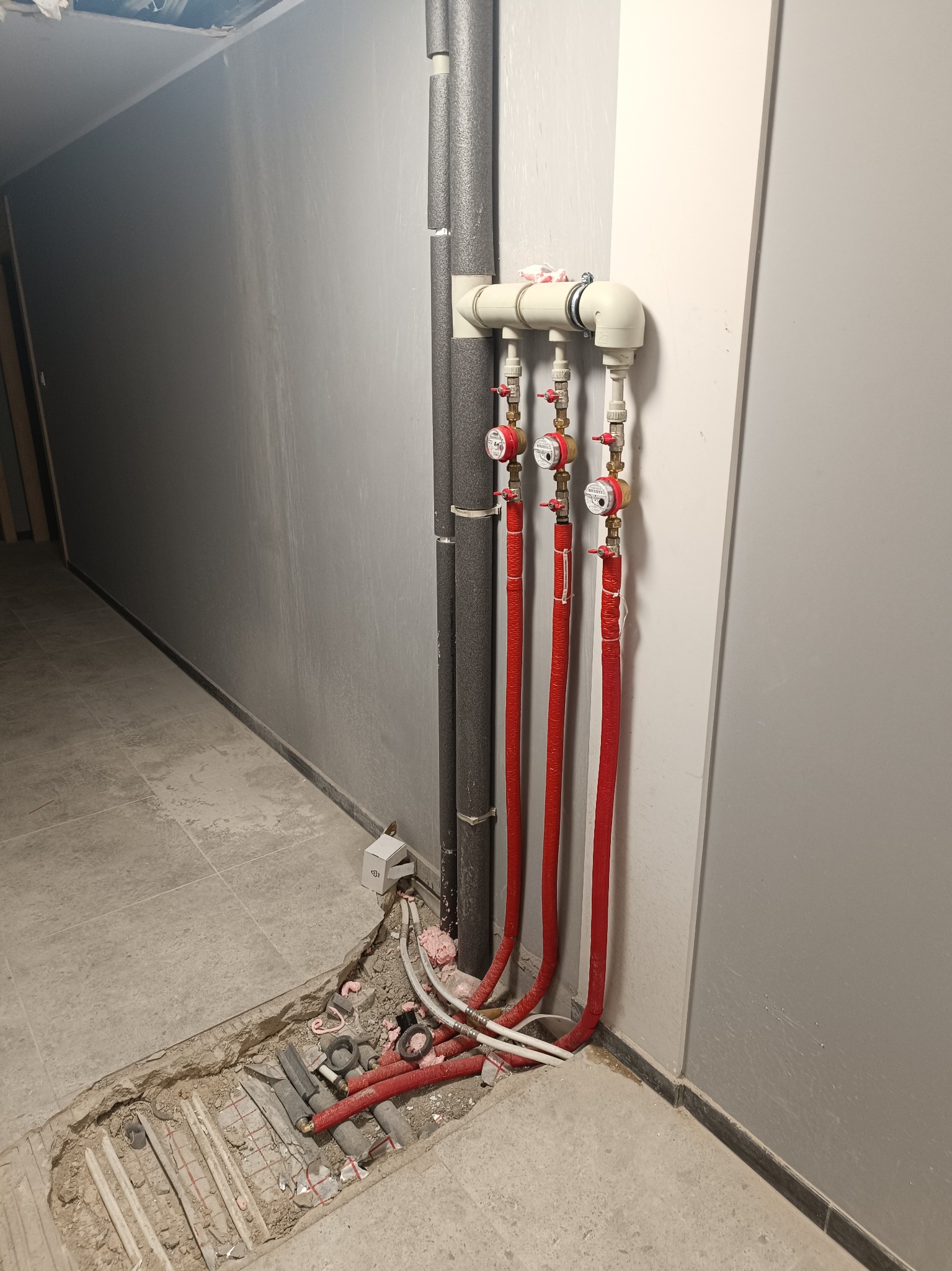 Rozdzielacz z czerwonymi rurami w korytarzu, widoczne liczniki i izolacja termiczna. Częściowo odkryta instalacja w podłodze, szare ściany i płytki podłogowe.