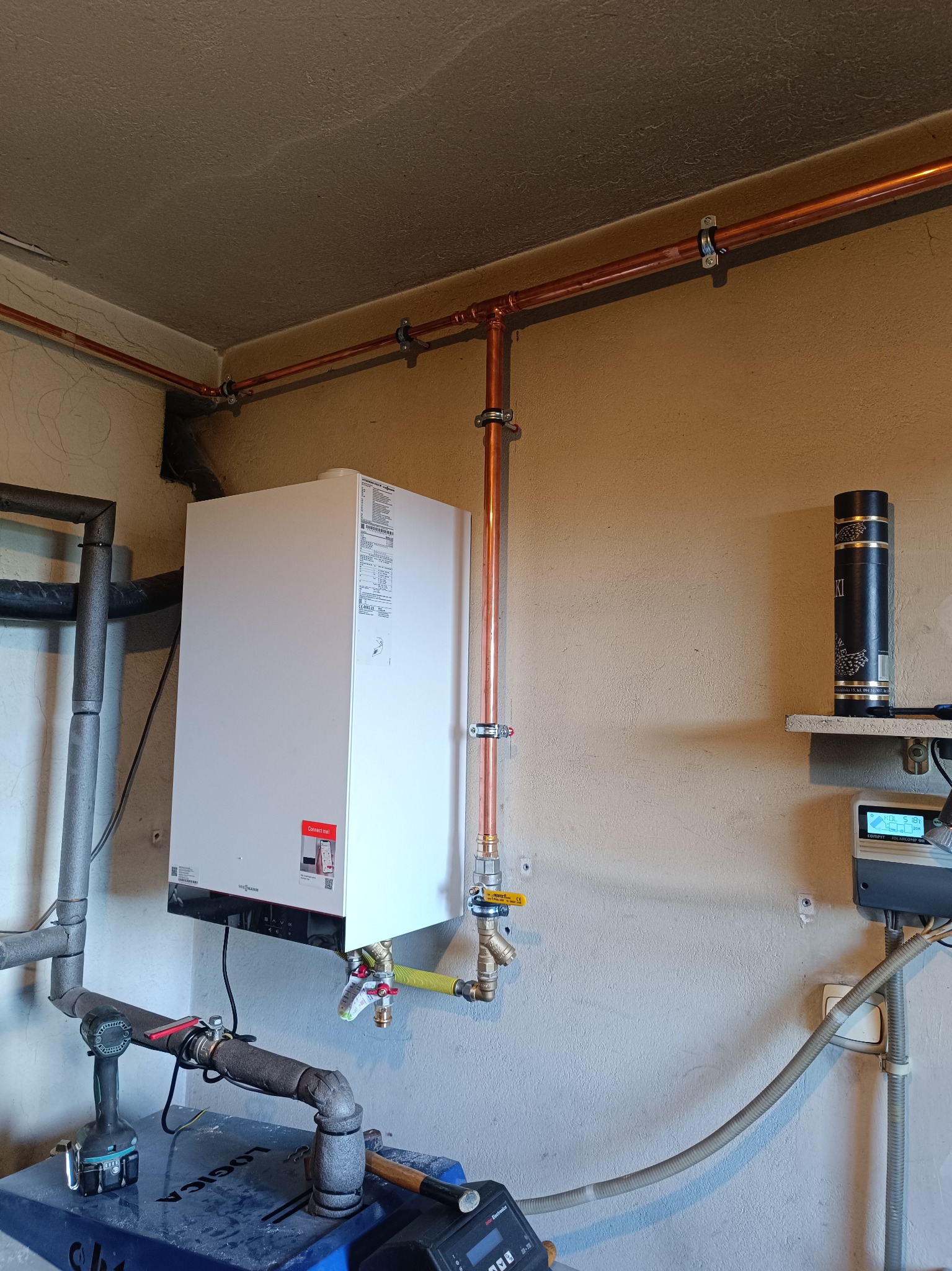 Biały piec gazowy Viessmann z miedzianą instalacją, zamontowany na beżowej ścianie. Widoczne rury, zawory i narzędzia. Ujęcie z dołu, oświetlenie naturalne.