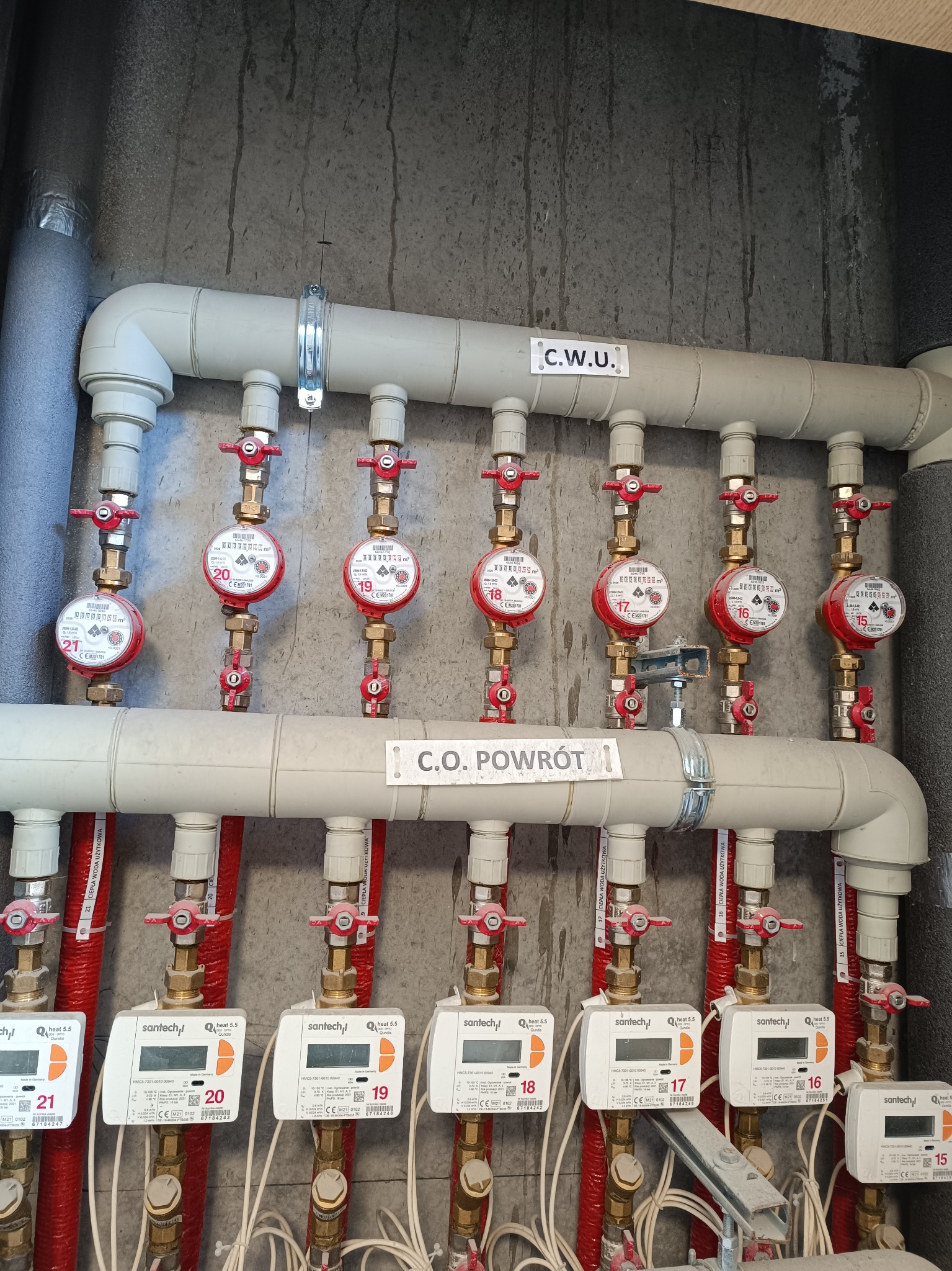 Rozdzielacz C.O. i C.W.U. z licznikami ciepła i wody użytkowej. Instalacja grzewcza z rurami w izolacji. Widoczne zawory i oznaczenia.
