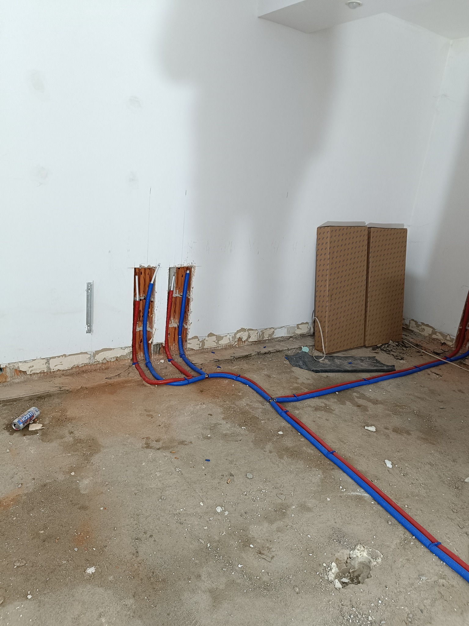 Wnętrze w trakcie prac hydraulicznych: czerwono-niebieskie rury prowadzone w bruzdach ściennych, podłoga w stanie surowym, kartony w rogu. Widok na instalację.
