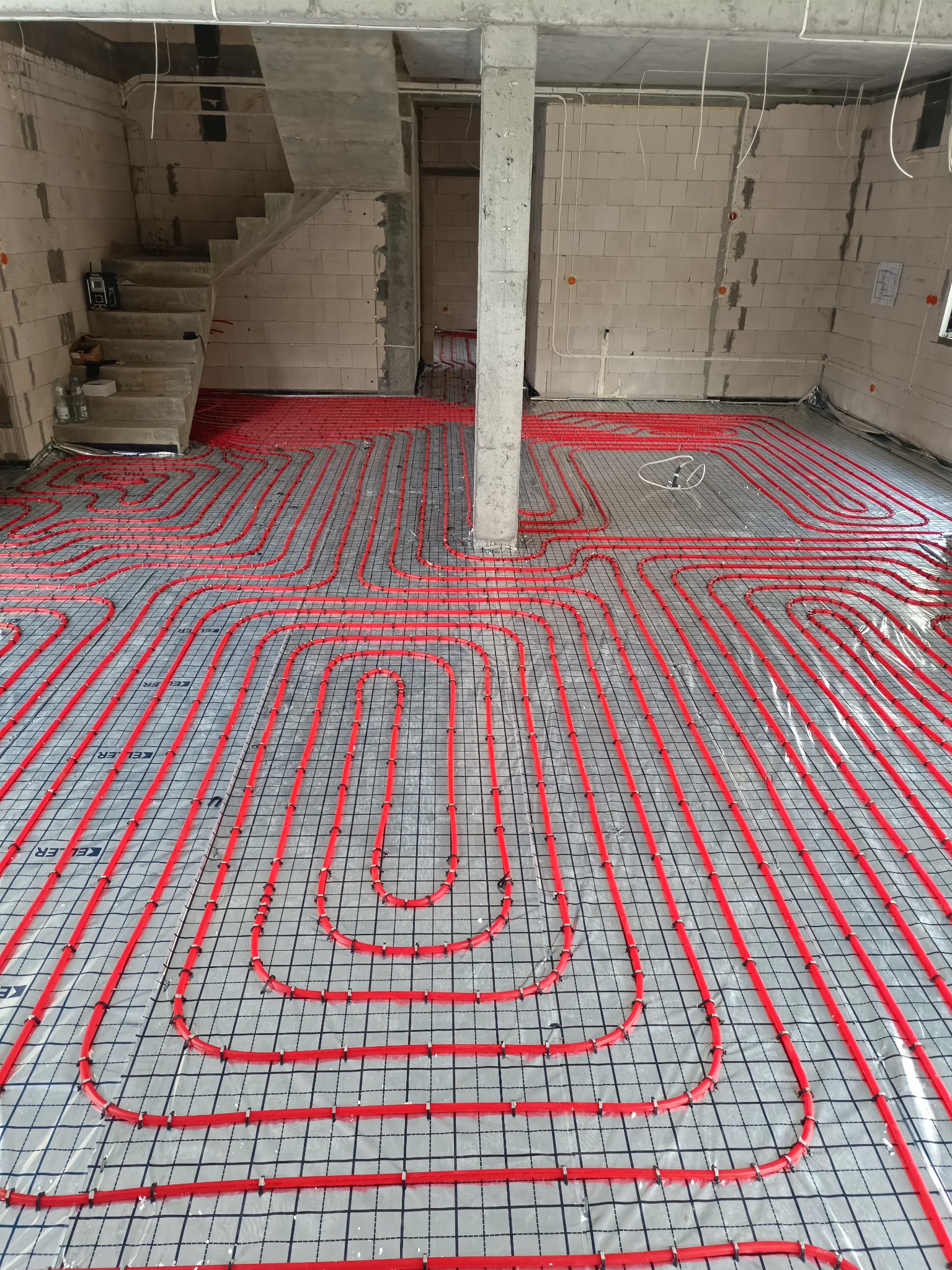 Czerwone rury ogrzewania podłogowego ułożone na folii z rastrem, widoczne elementy konstrukcyjne budynku w surowym stanie.