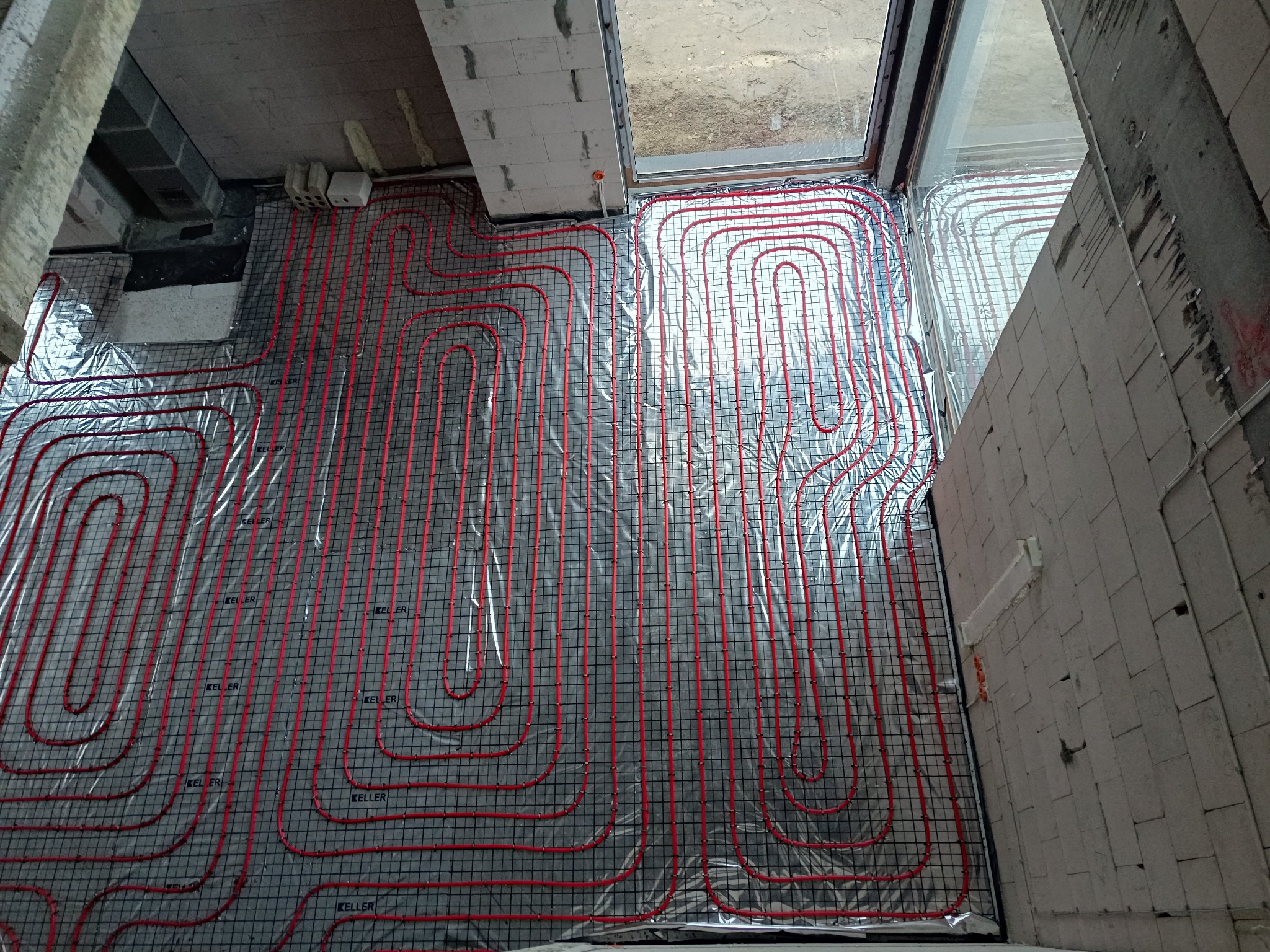 Czerwone rury ogrzewania podłogowego ułożone na folii odblaskowej, zamocowane do czarnej siatki. Widok z góry na instalację w surowym wnętrzu budynku.