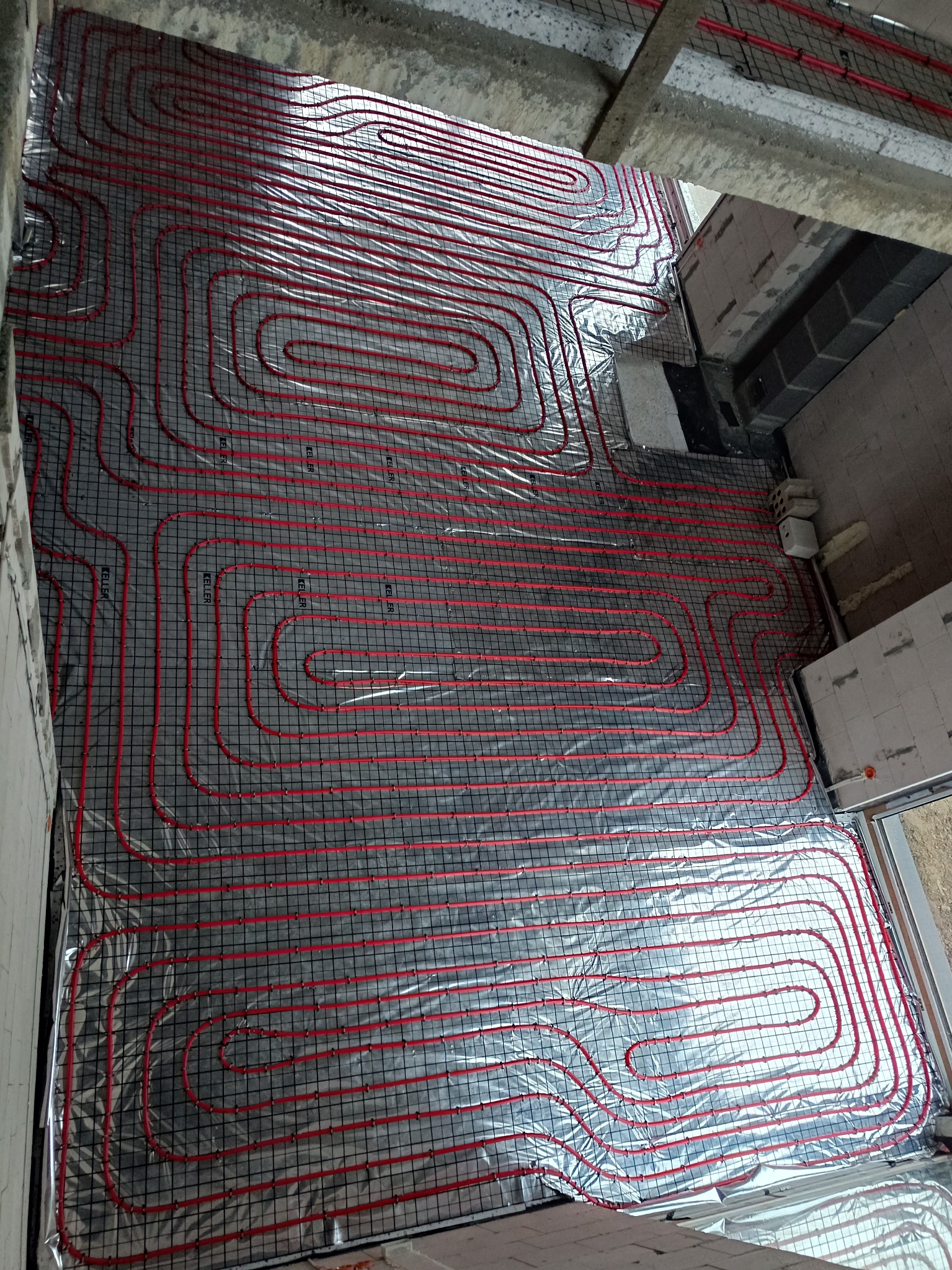 Czerwone rury ogrzewania podłogowego ułożone na srebrnej folii, zamocowane do czarnej siatki na podłodze w surowym wnętrzu budynku w trakcie budowy.