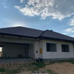 Surowy stan domu jednorodzinnego z ciemną dachówką na tle błękitnego nieba z chmurami, widoczne pustaki i otwory okienne bez ram.