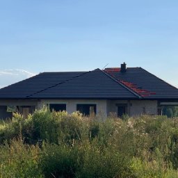Sztuka Budowania Sp. z o.o. - Surowy budynek jednorodzinny z ciemnym dachem w trakcie budowy, widok z poziomu wysokiej trawy, częściowo położona dachówka.