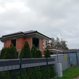 Sztuka Budowania Sp. z o.o. - Surowy stan domu z czerwonej cegły pod ciemnym dachem, widoczny zza ogrodzenia i żywopłotu z tuj, z kontenerem w tle.