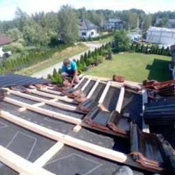 Pracownik w niebieskiej koszulce układa dachówkę na drewnianej konstrukcji dachu, widok z góry na zielony ogród i domy w tle.