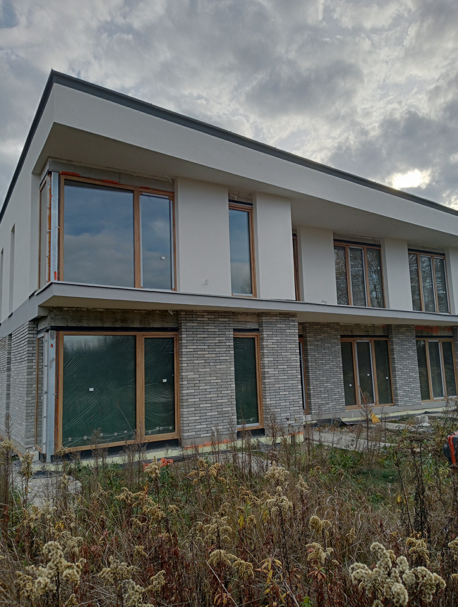 Dwukondygnacyjny dom w trakcie budowy w Warszawie, z szarej cegły na parterze i białym tynkiem na piętrze, drewniane ramy okienne, okna zabezpieczone folią, widoczne chwasty na pierwszym planie.