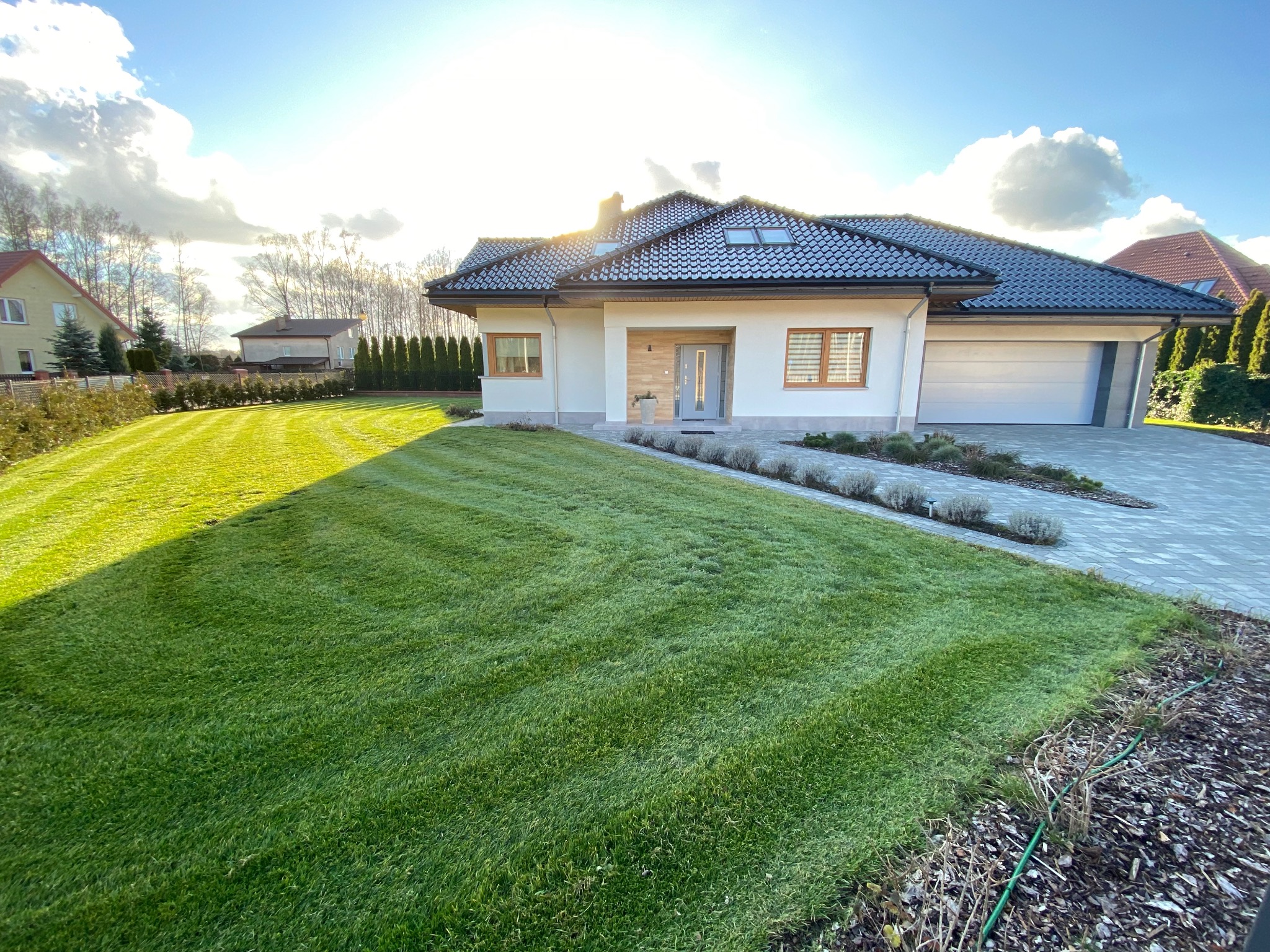 Nowoczesny dom jednorodzinny z garażem wbudowanym, z zadbanym trawnikiem z wykoszonymi pasami, oświetlony popołudniowym słońcem.