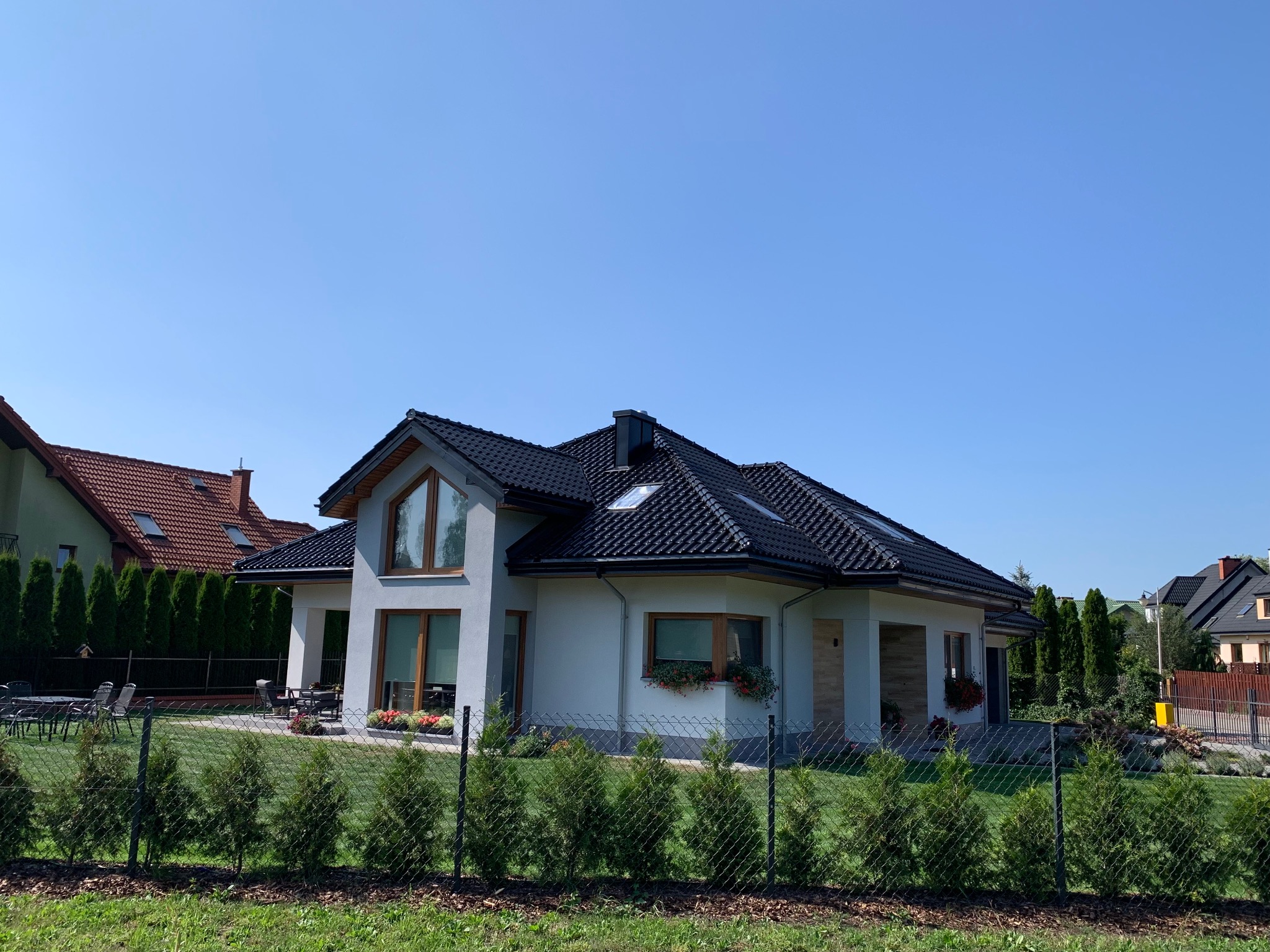 Nowoczesny dom jednorodzinny z ciemną dachówką, jasną elewacją i dużymi oknami, otoczony zadbanym trawnikiem i niskim żywopłotem z iglaków.