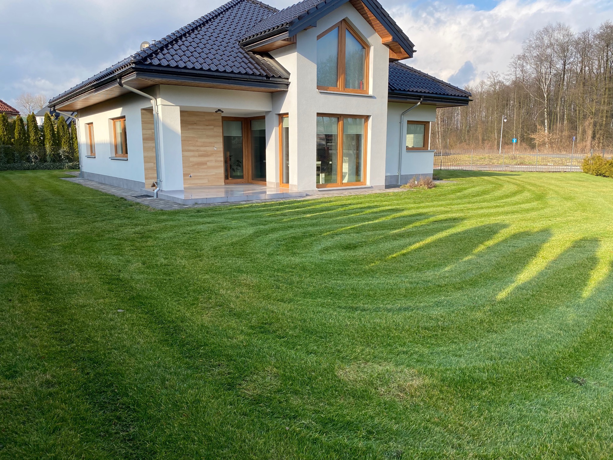 Nowoczesny dom jednorodzinny z białymi ścianami, ciemną dachówką i drewnianymi ramami okiennymi, otoczony równo przystrzyżonym trawnikiem z widocznymi śladami kosiarki.