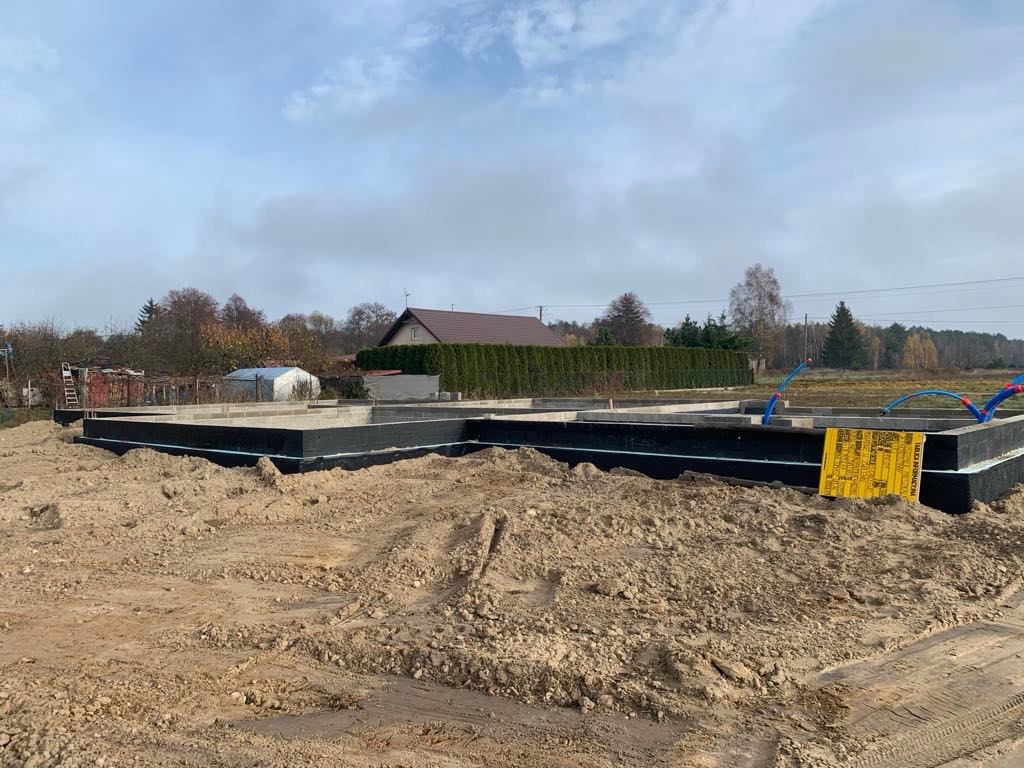 Wylany fundament z izolacją przeciwwilgociową i wystającymi rurami instalacji wodnej, przygotowany do dalszych prac budowlanych na działce z widocznymi drzewami w tle.