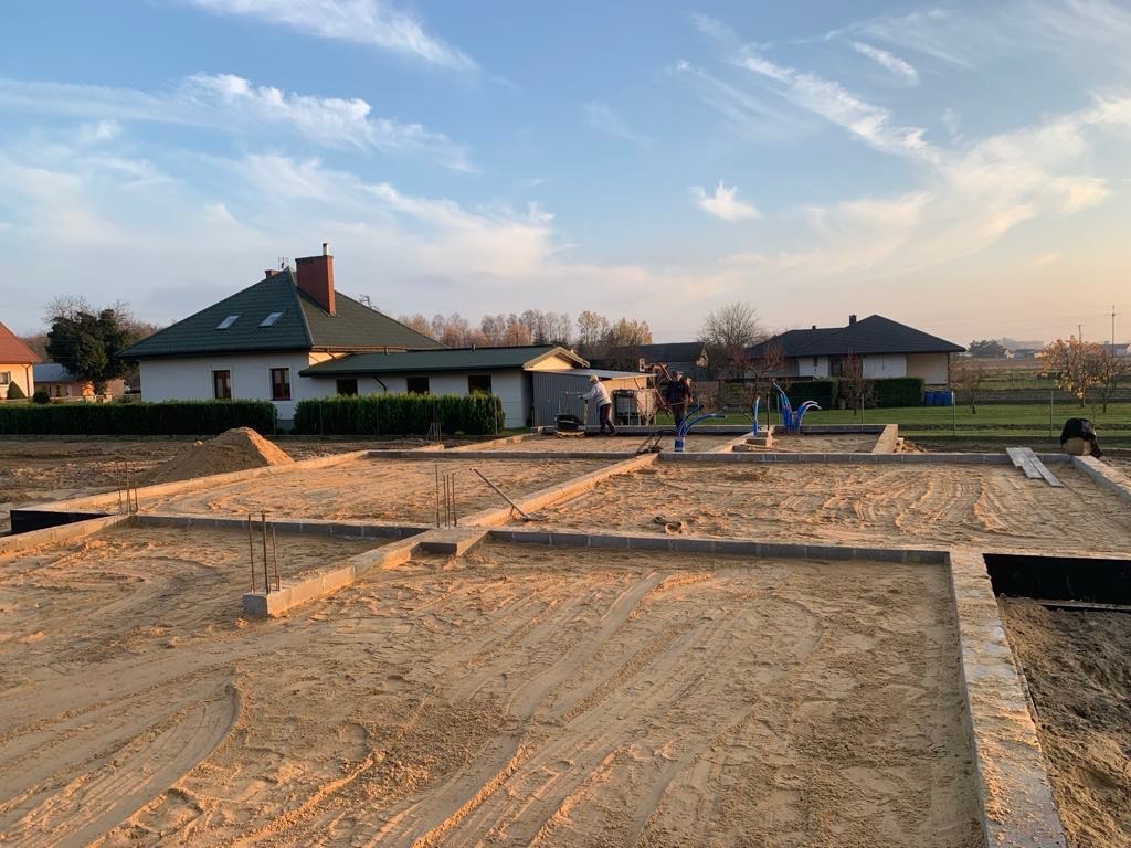 Fundamenty domu w trakcie budowy, widoczne zbrojenia i bloczki fundamentowe, dwóch pracowników w tle, krajobraz wiejski.