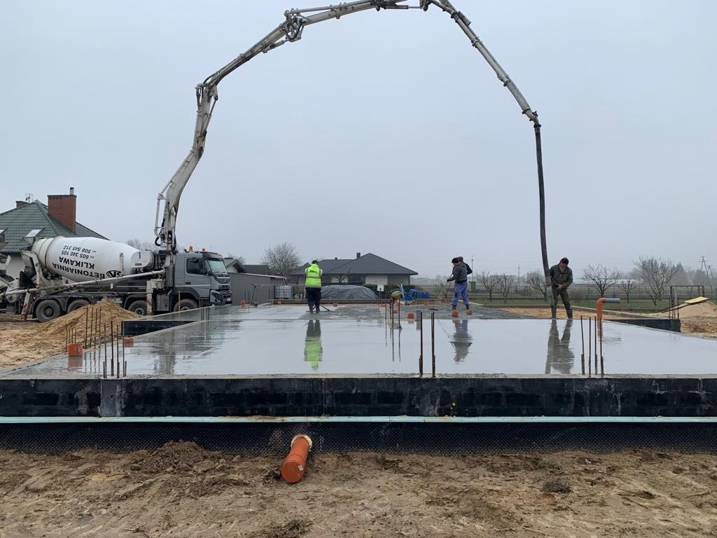 Wylewanie fundamentów żelbetowych pod budynek jednorodzinny za pomocą pompy do betonu. Widoczni pracownicy i betonowóz z logo betoniarni.