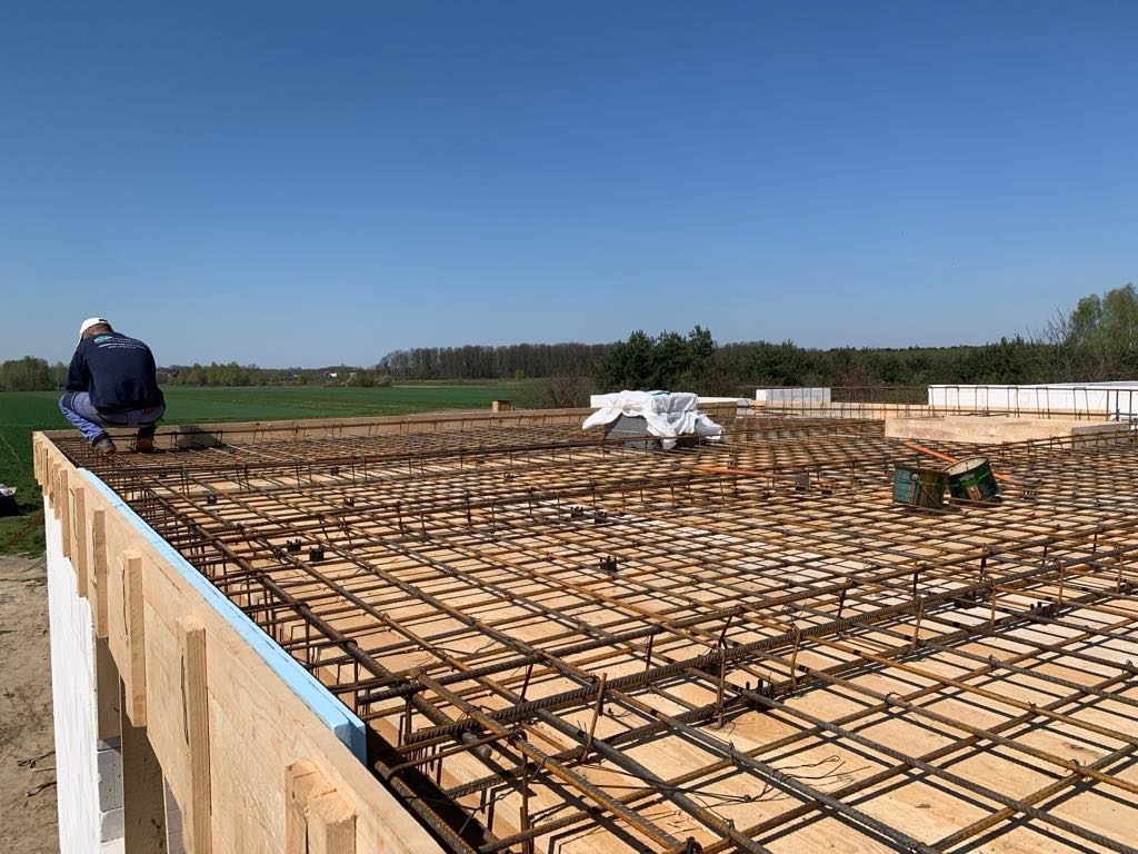 Prace zbrojarskie na budowie, widok z góry na siatkę zbrojeniową na deskowaniu, pracownik w kasku w tle, błękitne niebo.