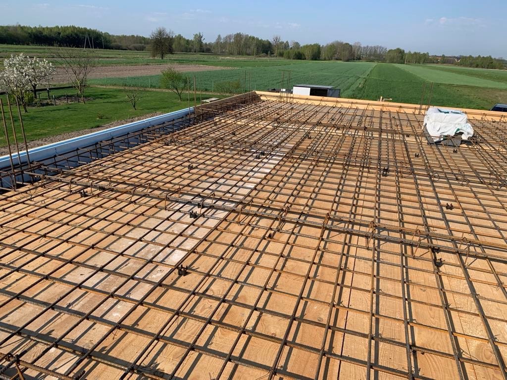 Zbrojenie płyty stropowej na deskowaniu, widok z góry na siatkę z prętów stalowych i drewniane płyty, w tle zielone pola i drzewa.