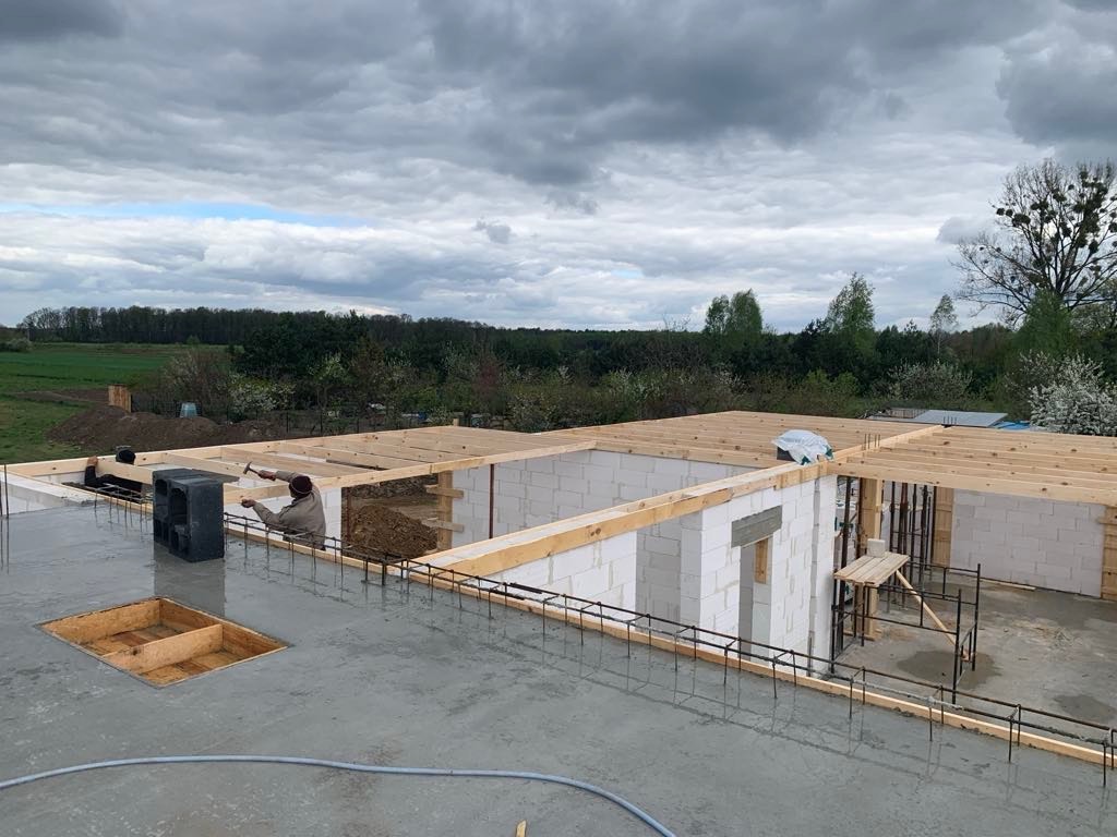 Wznoszenie ścian z bloczków betonowych na świeżo wylanym stropie, widoczne zbrojenia i drewniane szalunki, pracownik z młotkiem w tle, pochmurne niebo.