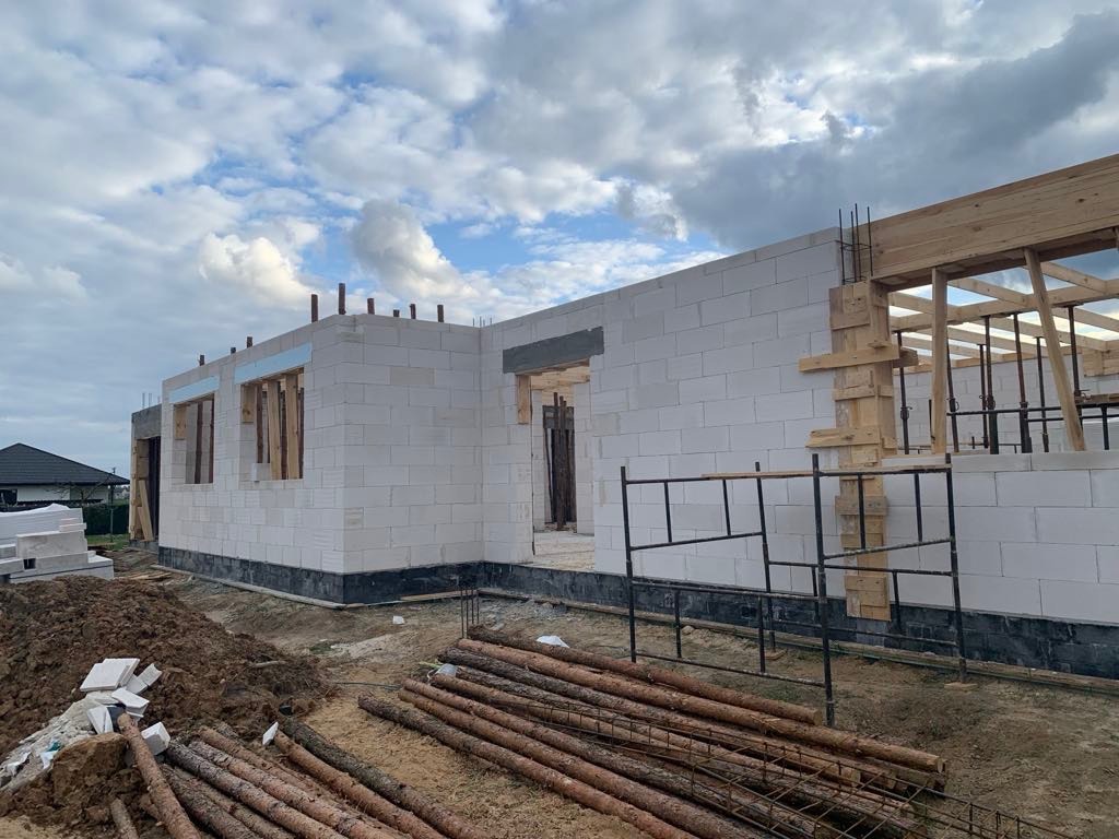 Wznoszenie ścian parterowego budynku z białych bloczków, widoczne drewniane szalunki okienne i drzwiowe, oraz konstrukcja dachu w trakcie budowy, na pierwszym planie stos drewna i ziemi.