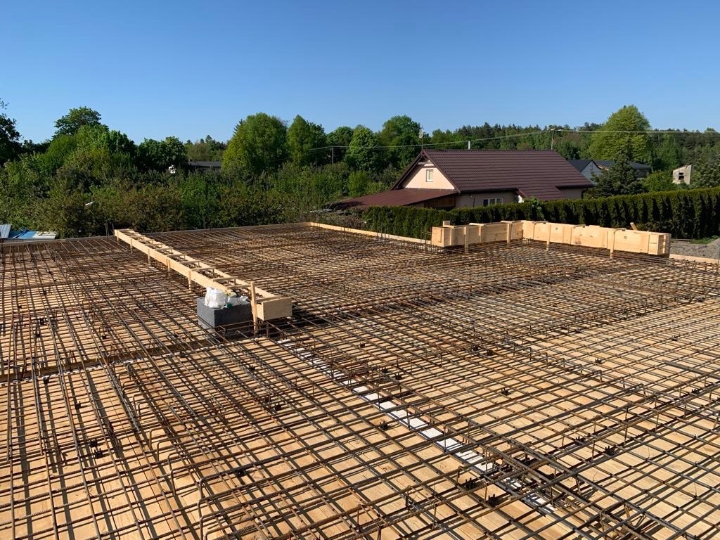Zbrojenie stropu budynku w trakcie budowy, widoczne drewniane szalunki i siatka z prętów stalowych, w tle dom jednorodzinny z brązowym dachem.